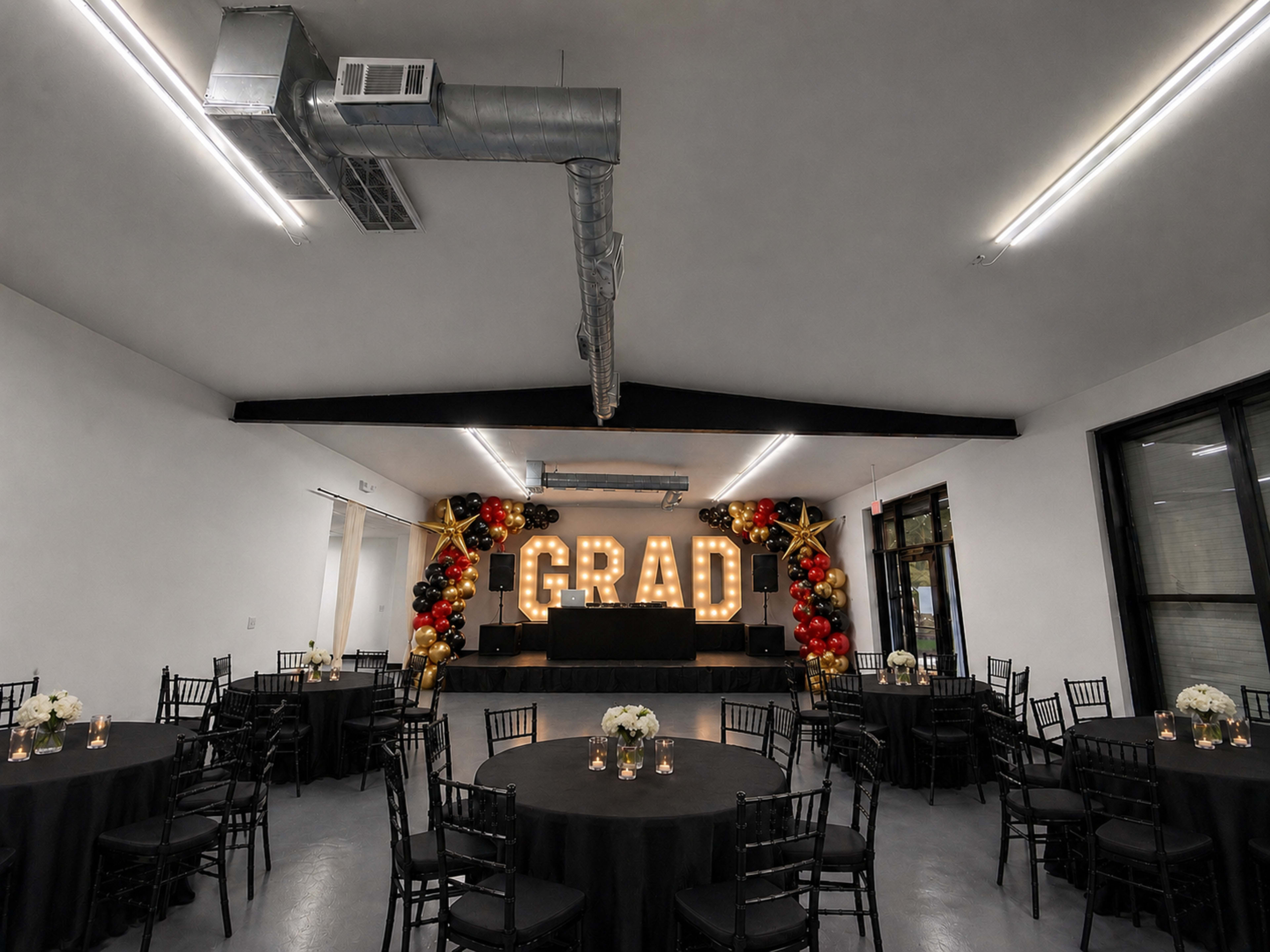 The image shows a decorated indoor space for a graduation celebration, featuring large illuminated letters spelling "GRAD" and tables arranged with black tablecloths and floral centerpieces.