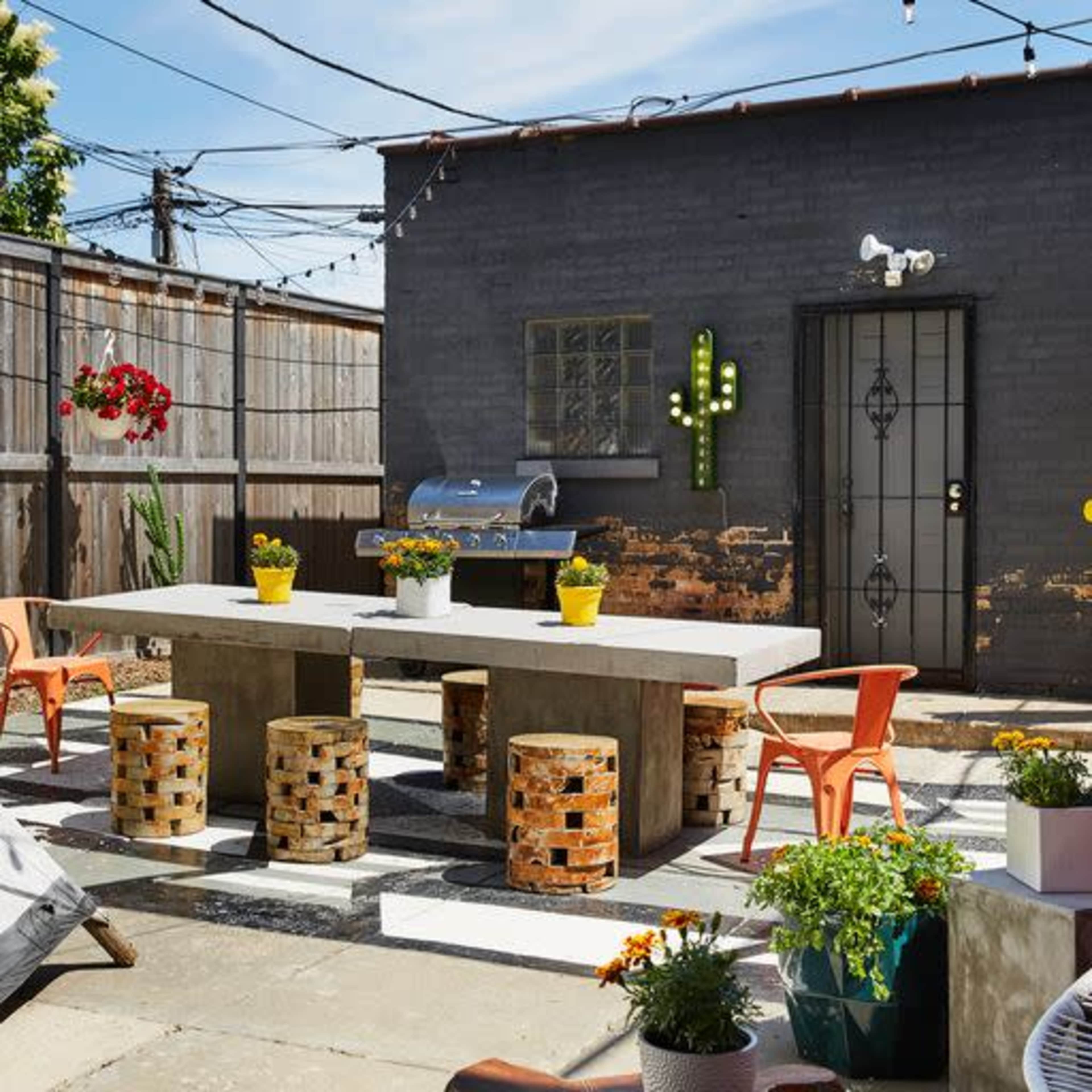 A modern outdoor patio features a concrete table surrounded by bright, patterned chairs, potted plants, and string lights overhead.