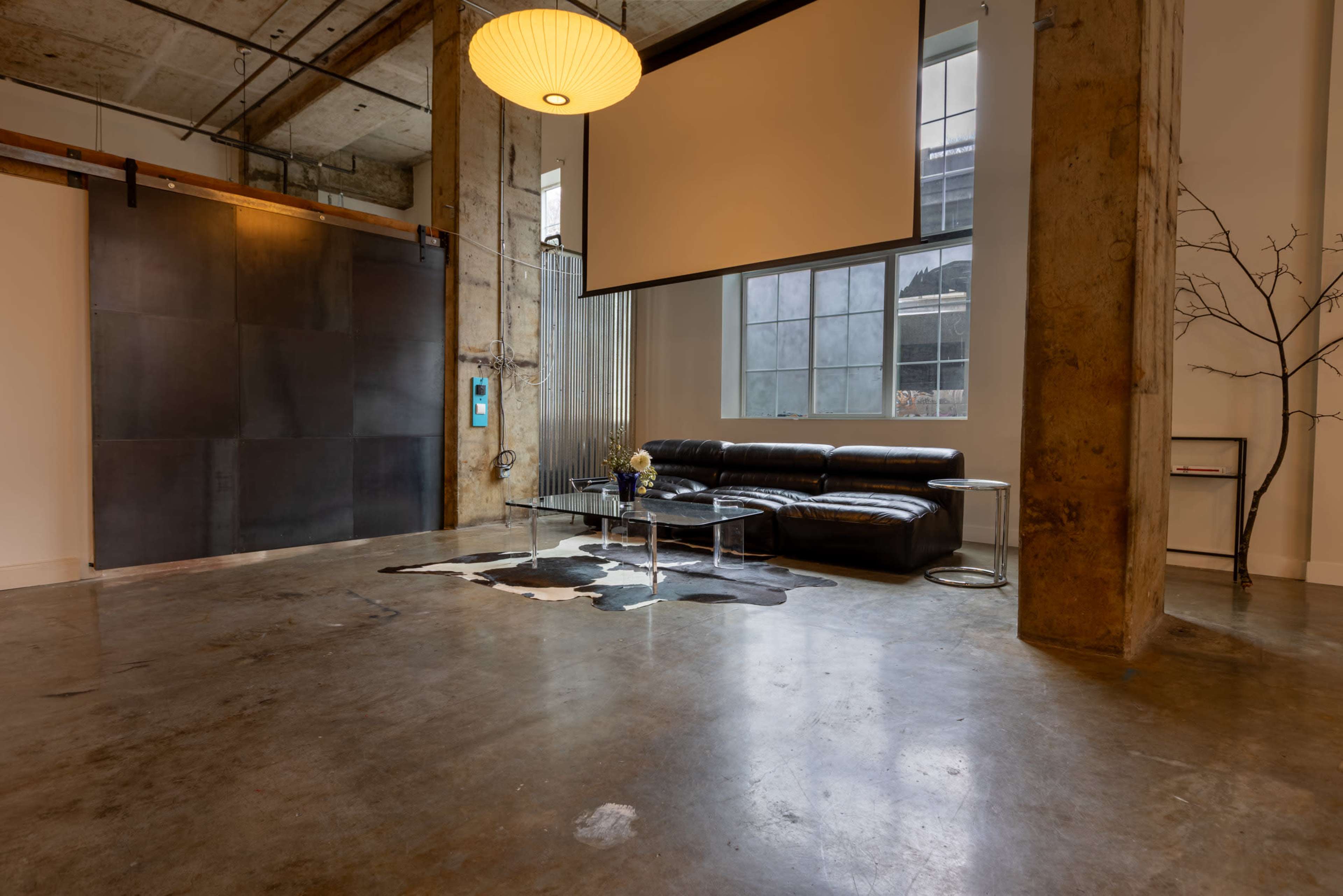 The image shows a modern loft-like interior with polished concrete floors, a black leather sofa, a large window, and a decorative cowhide rug.