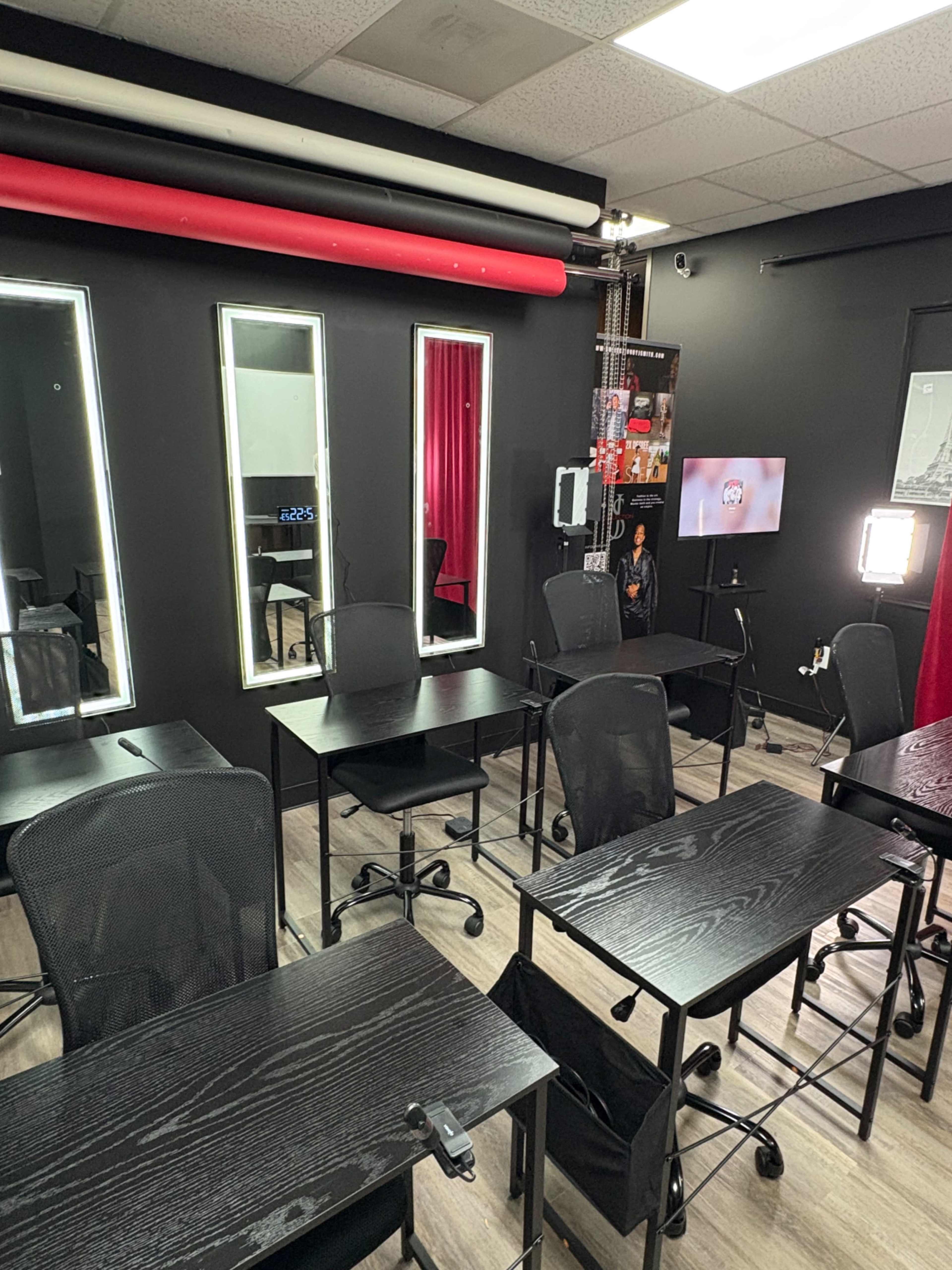 The image shows a classroom setup with several black desks and chairs arranged in a room with black walls, mirrors, and a wall-mounted screen.