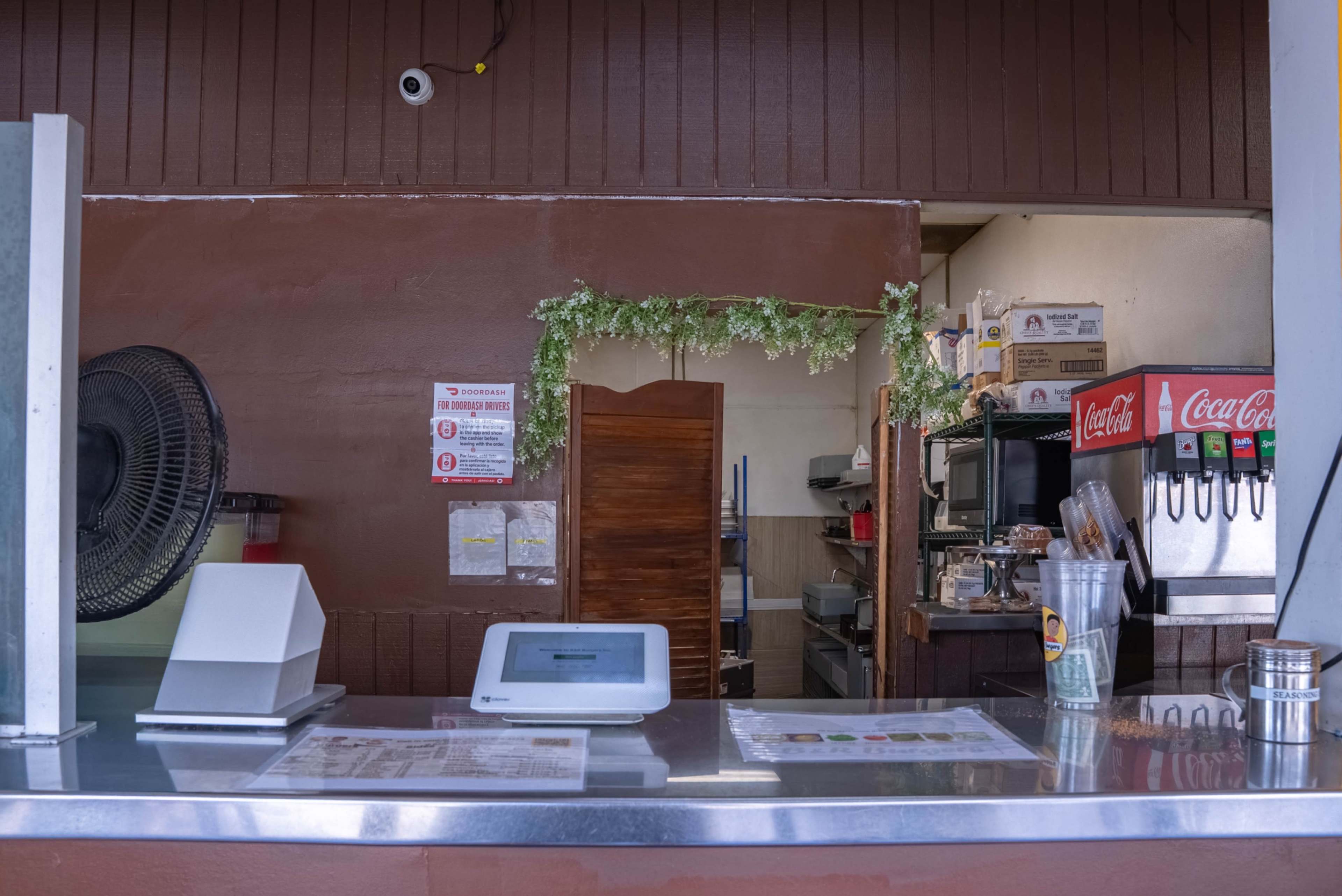 The image shows the interior of a small food establishment with a service counter, a fan on the wall, and kitchen equipment visible in the background.