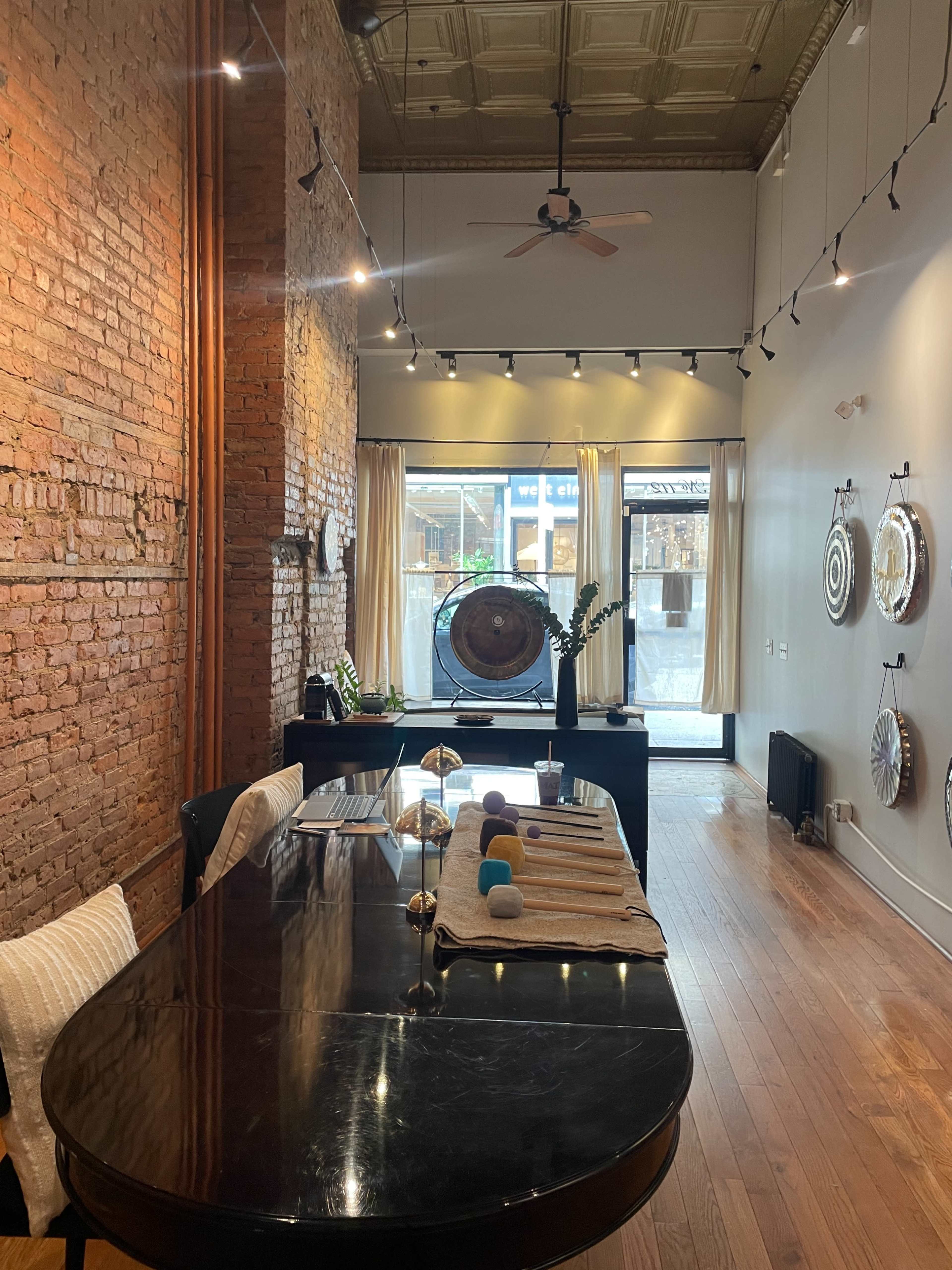 The image shows an interior space with exposed brick walls, wooden flooring, a large black table in the center, and various decorative items placed around the room, including a large window that lets in natural light.