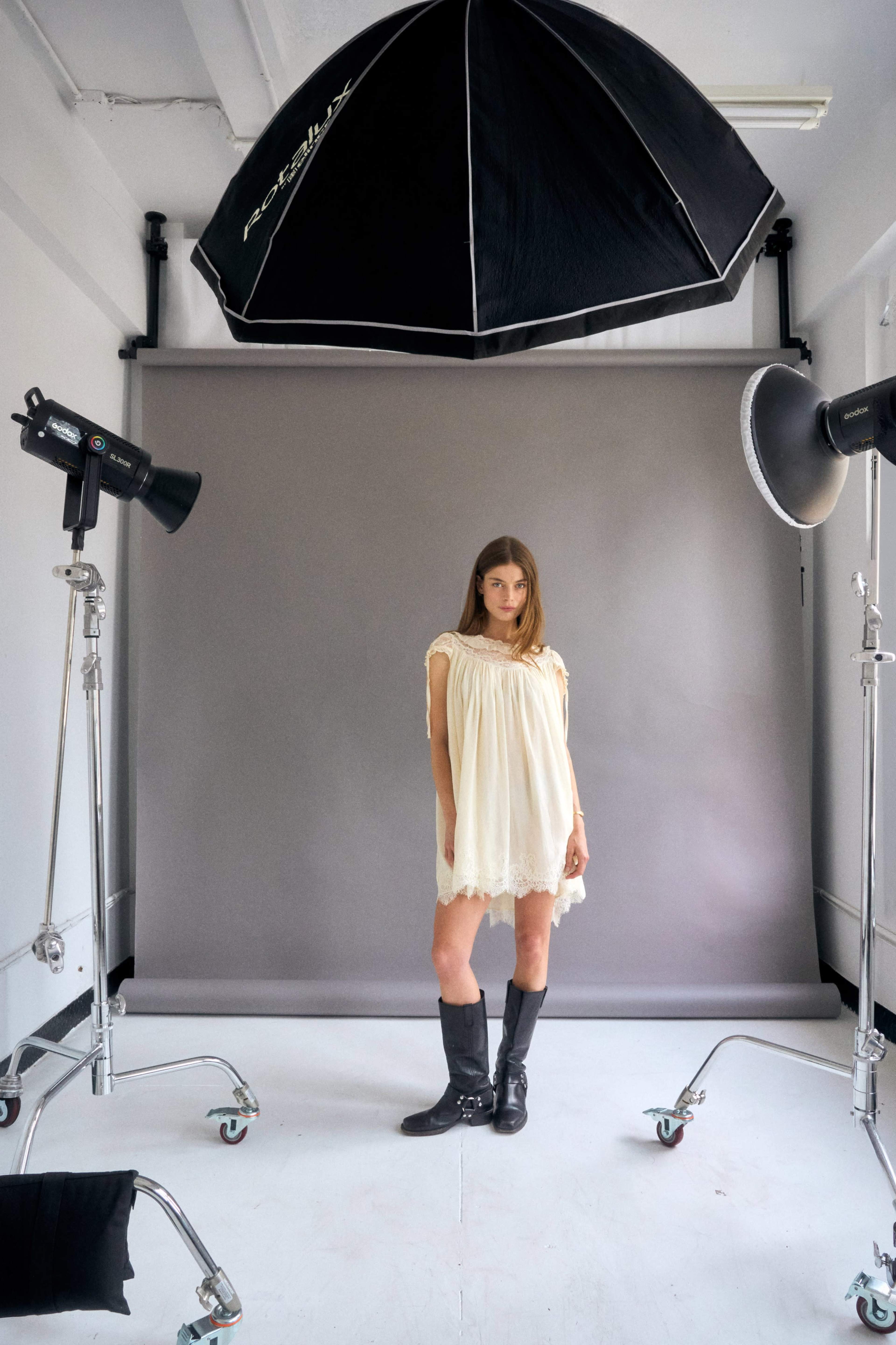 A model stands in a studio setting, wearing a light dress and black boots, with a backdrop and lighting equipment positioned around her.
