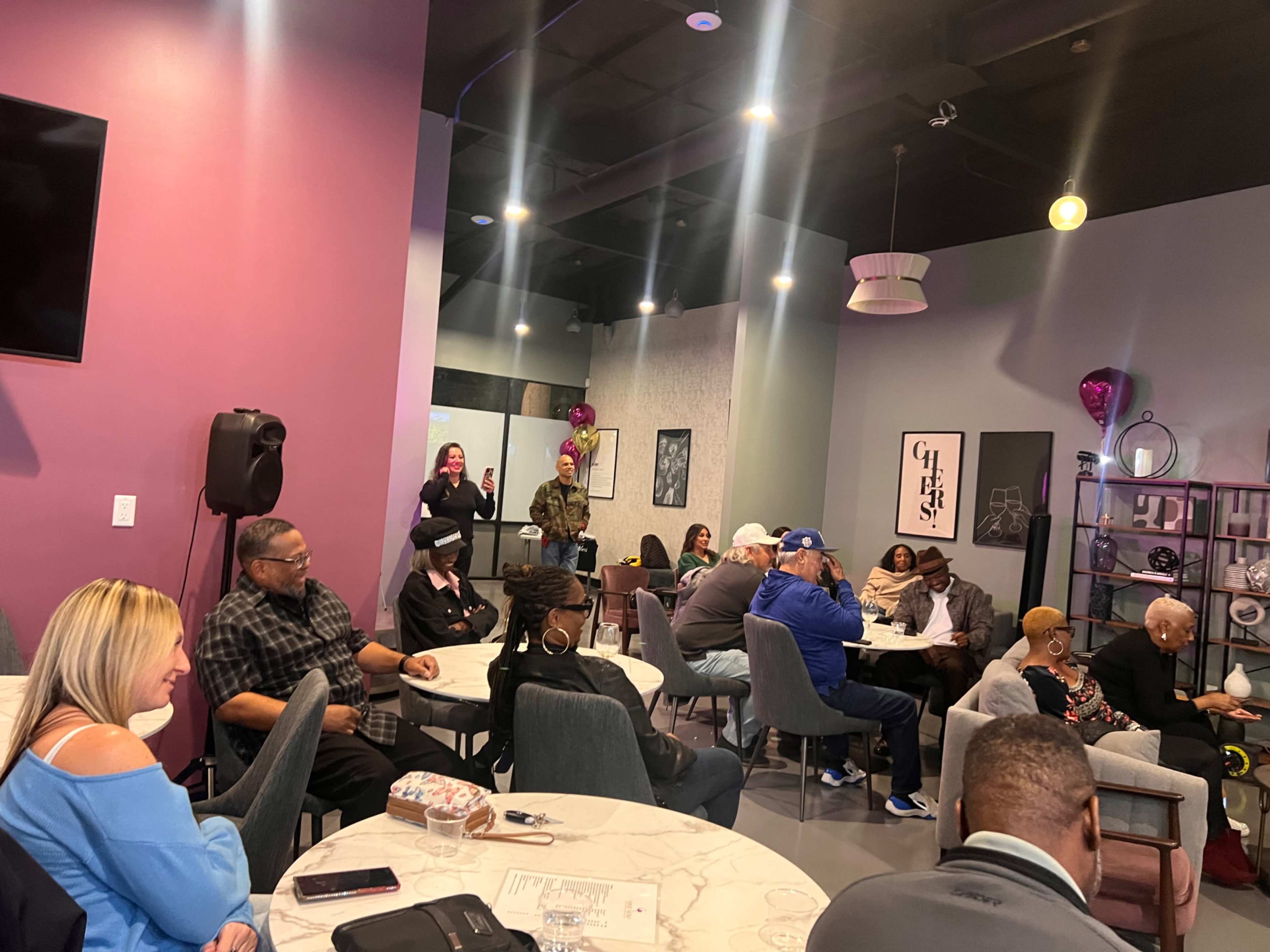 A diverse group of people is gathered in a stylish, modern venue, seated at tables and facing a speaker area.