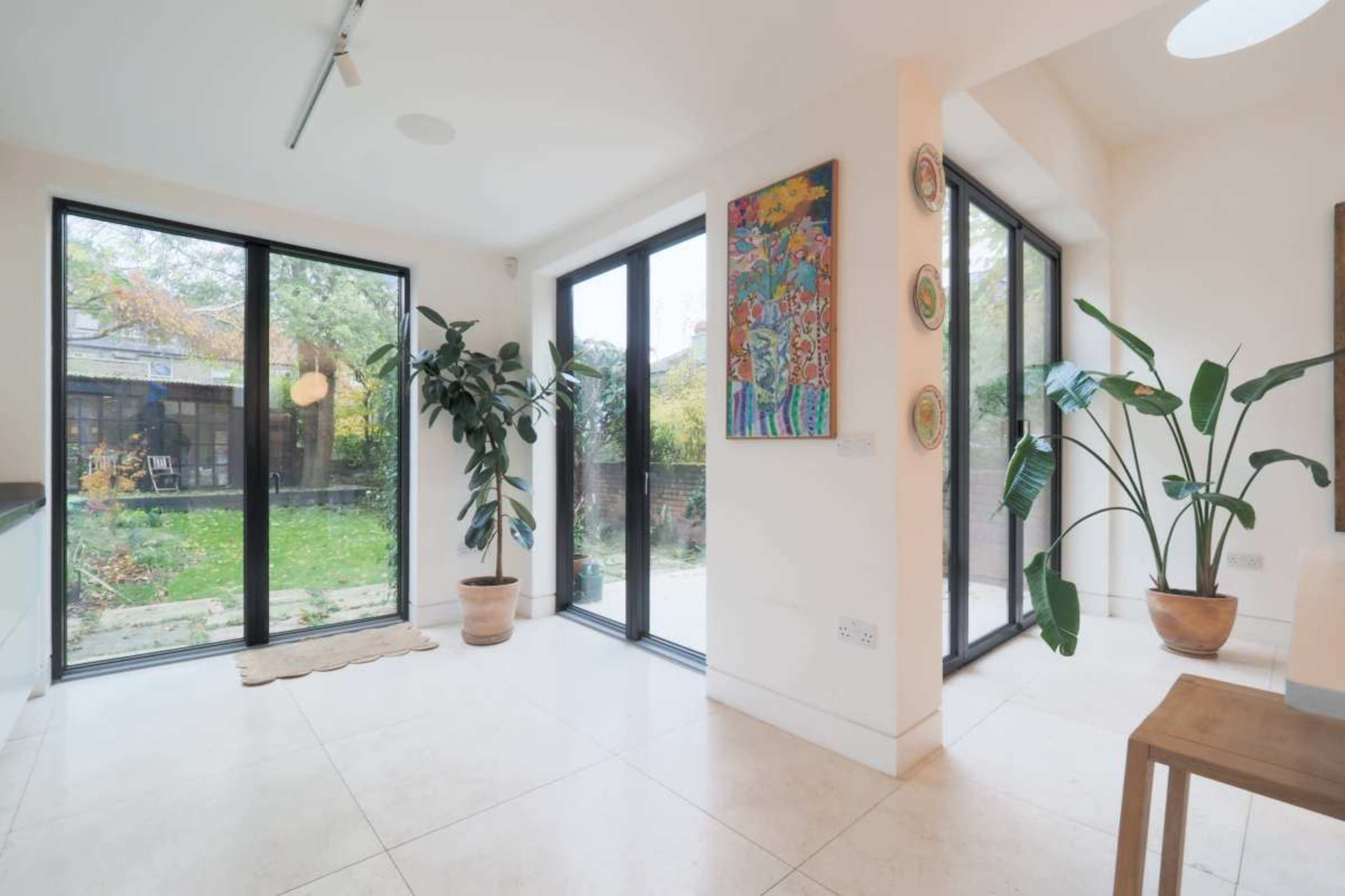 The image shows an interior room with large glass windows, a potted plant, and a view of a garden outside.