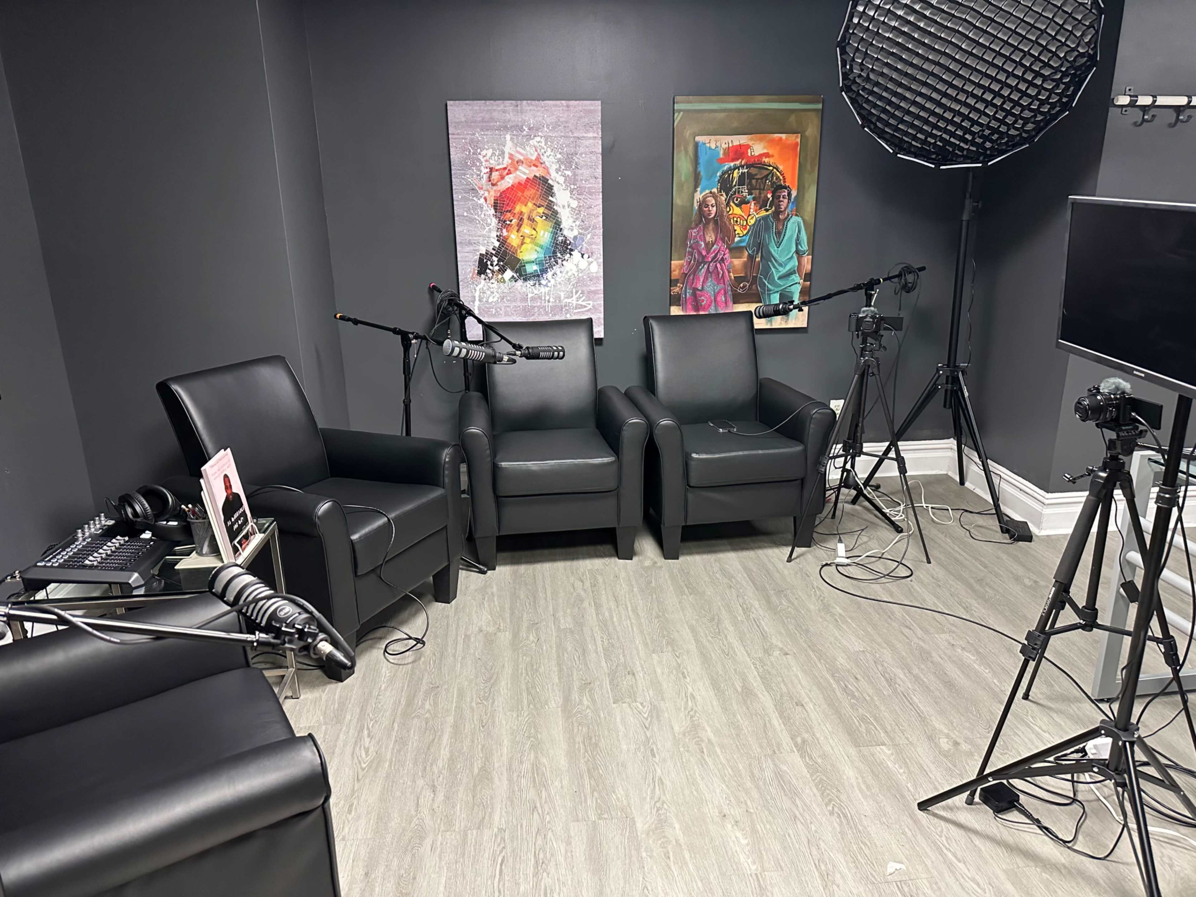 A recording studio setup with three leather chairs, two colorful wall art pieces, and several cameras and microphones arranged around the room.