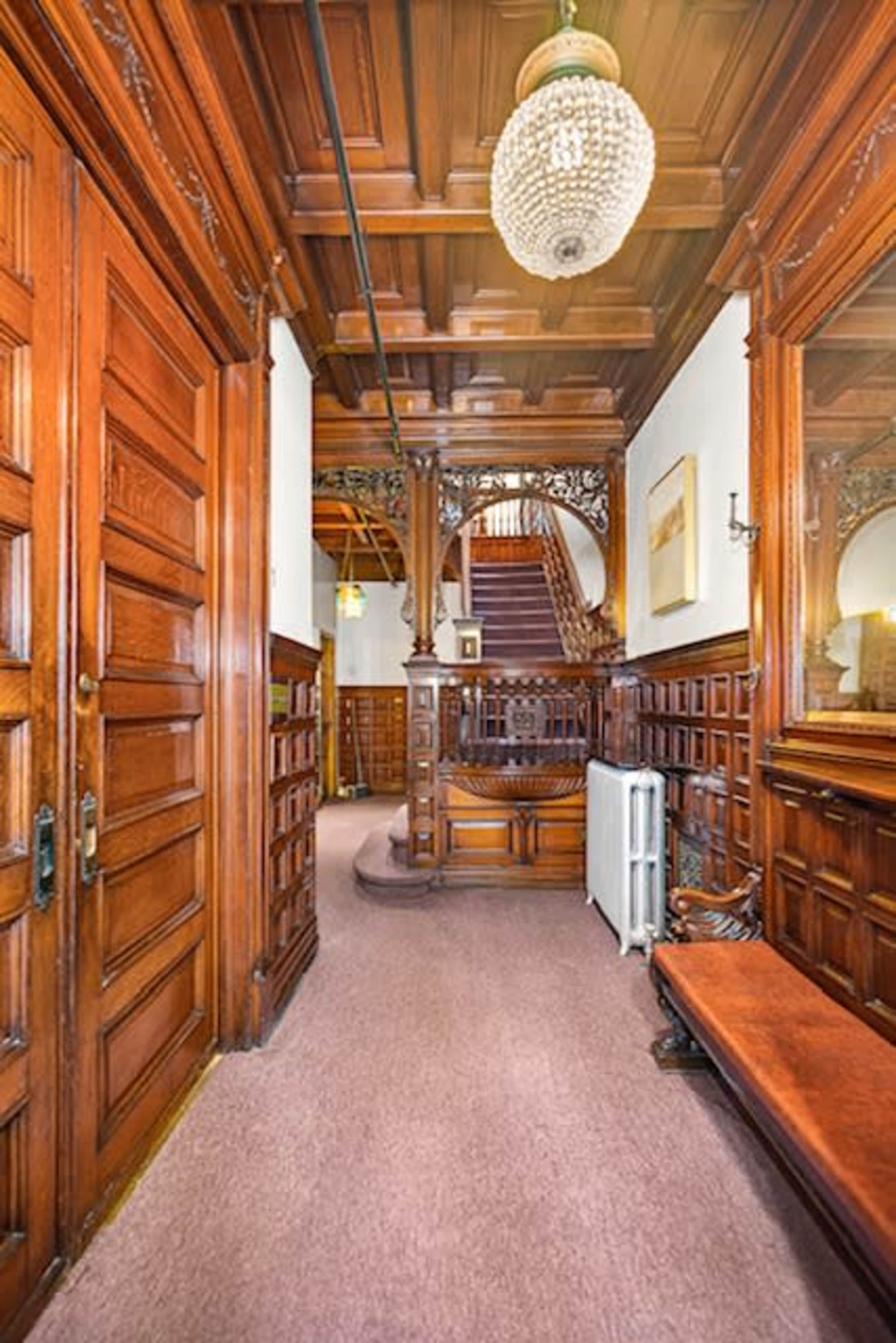 A wooden hallway with intricately carved paneling leads to a staircase, featuring a chandelier and a bench along one side.