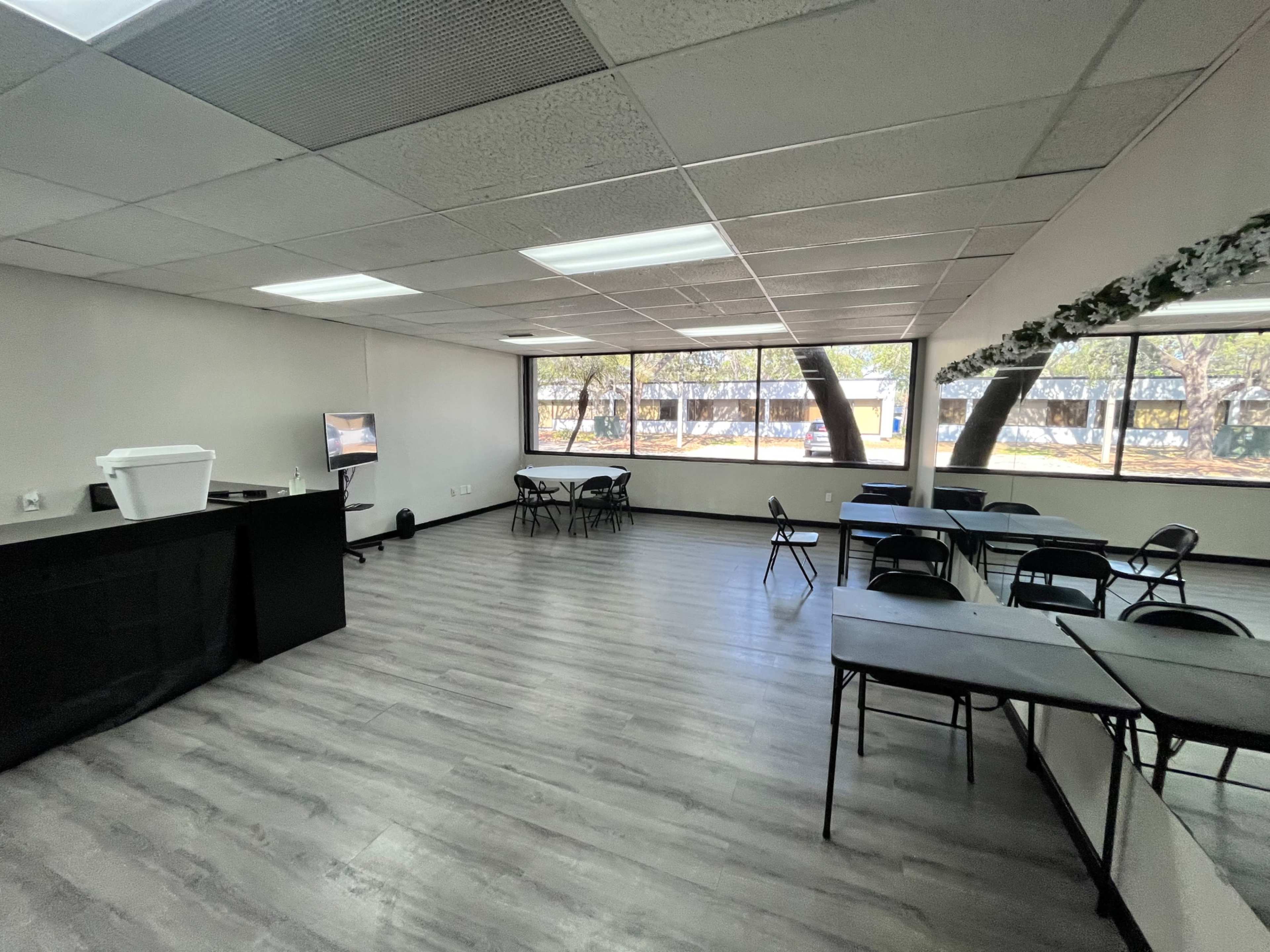 A spacious indoor room with several black chairs and tables arranged, large windows allowing natural light, and a small counter with a white container.