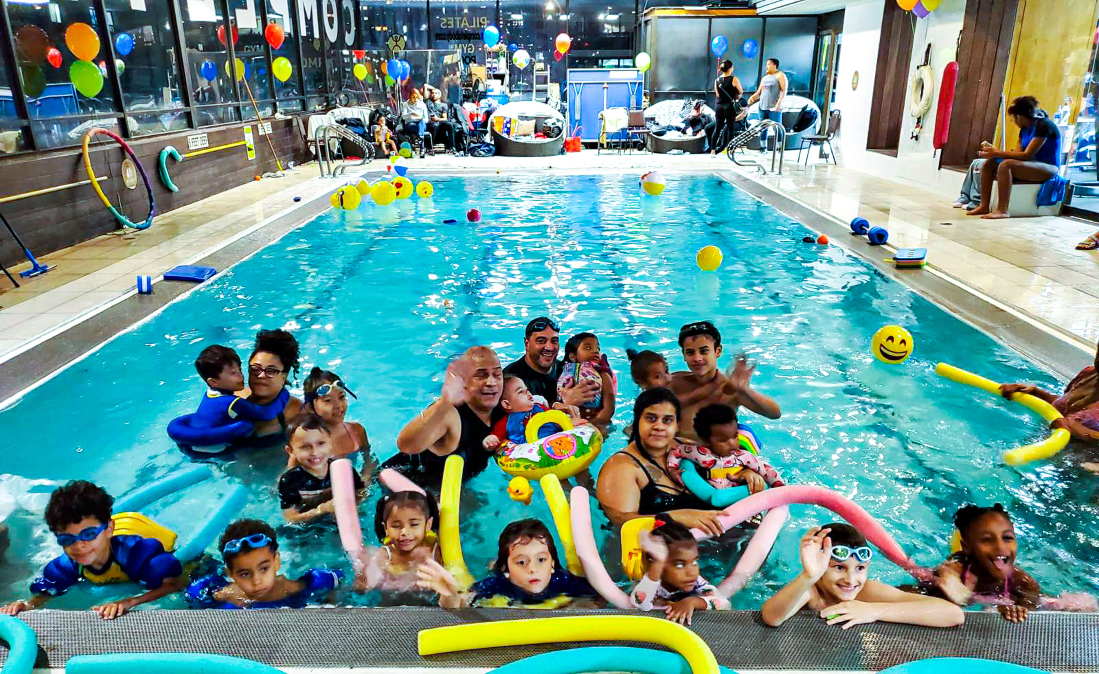 A large group of children and adults are playing in a pool filled with colorful inflatable toys and swim noodles.