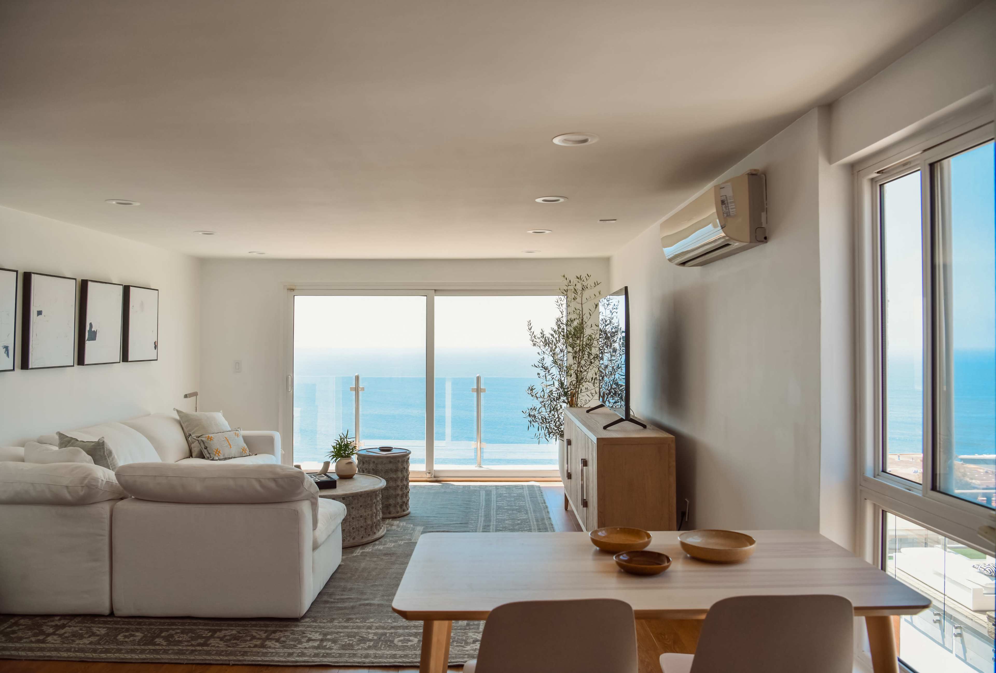 The image shows a bright living room with a large window overlooking the ocean, featuring a sofa, a coffee table, and a dining table set for four.