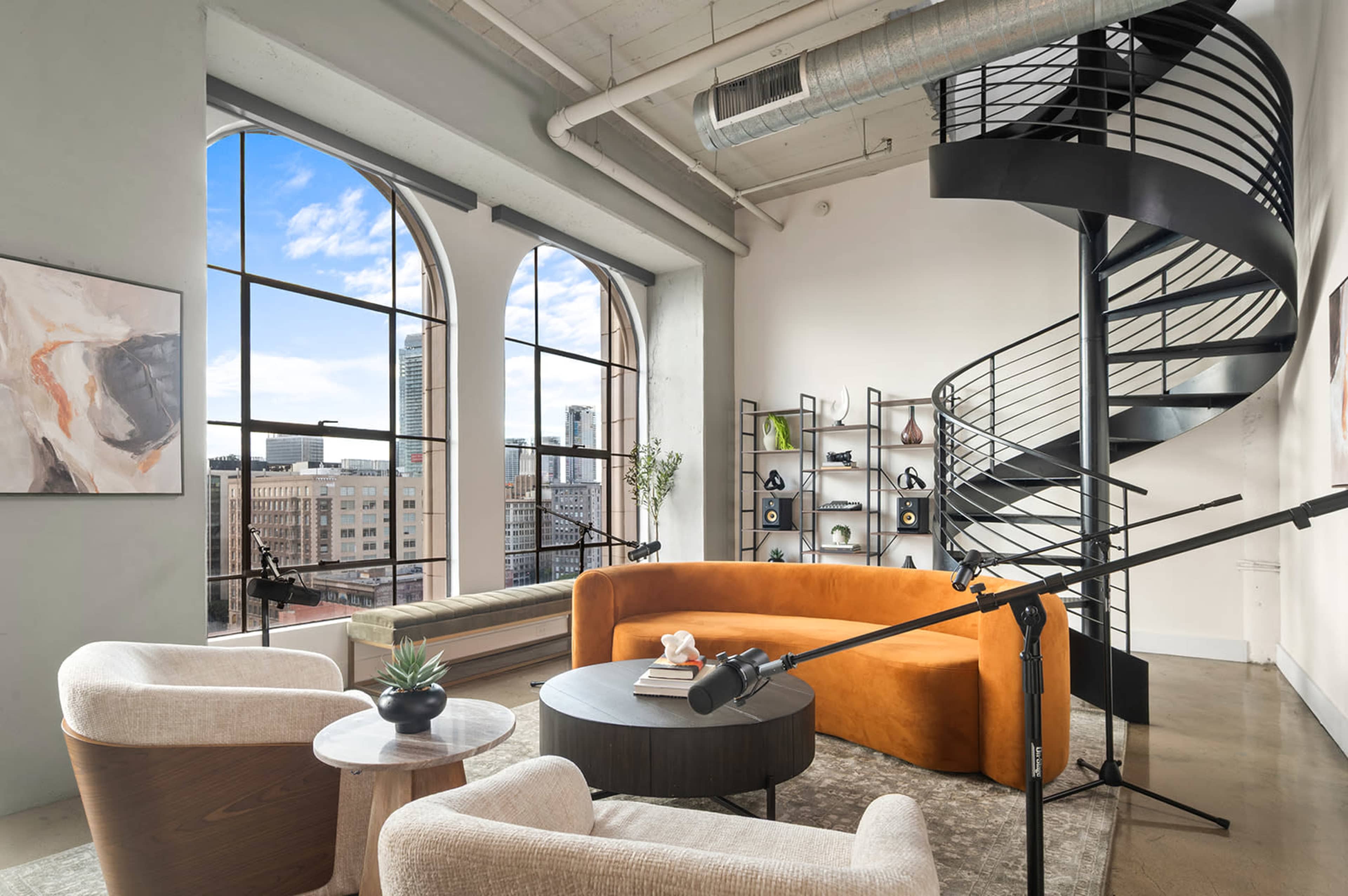 The image shows a modern living space with large arched windows, a spiral staircase, and contemporary furniture, including an orange sofa and two armchairs.