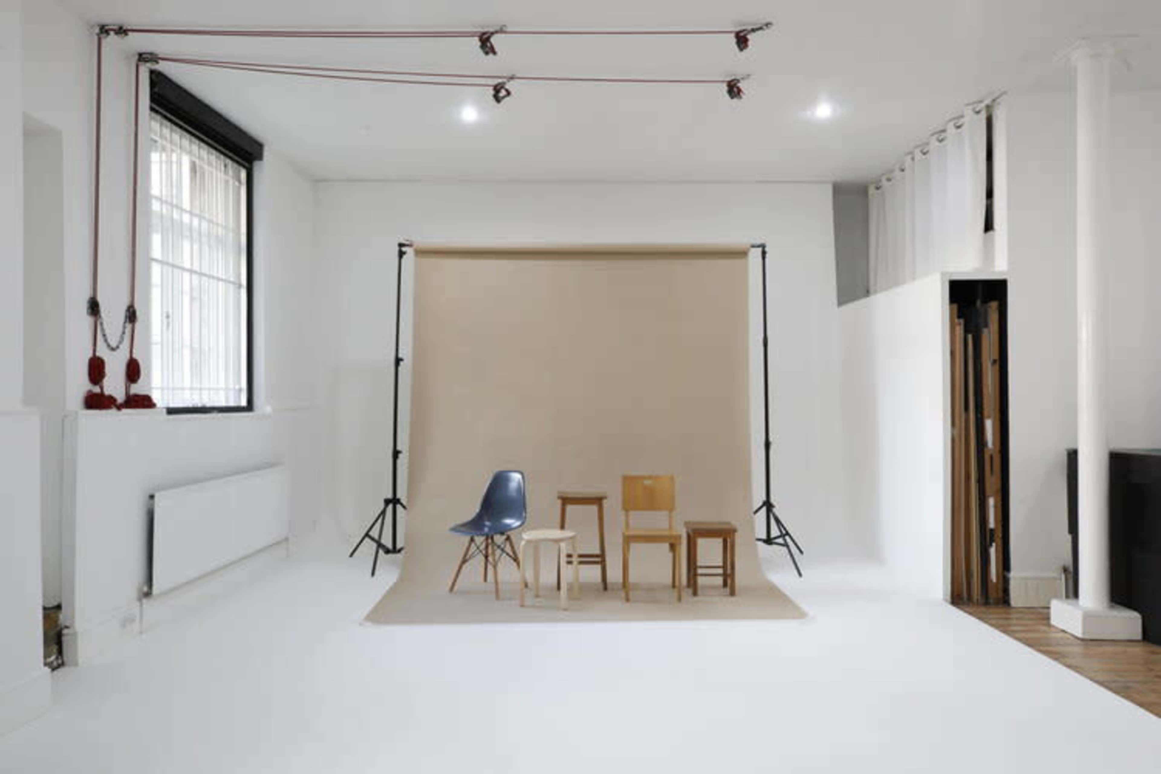 The image depicts a minimalistic studio space featuring three wooden chairs arranged in front of a beige backdrop.