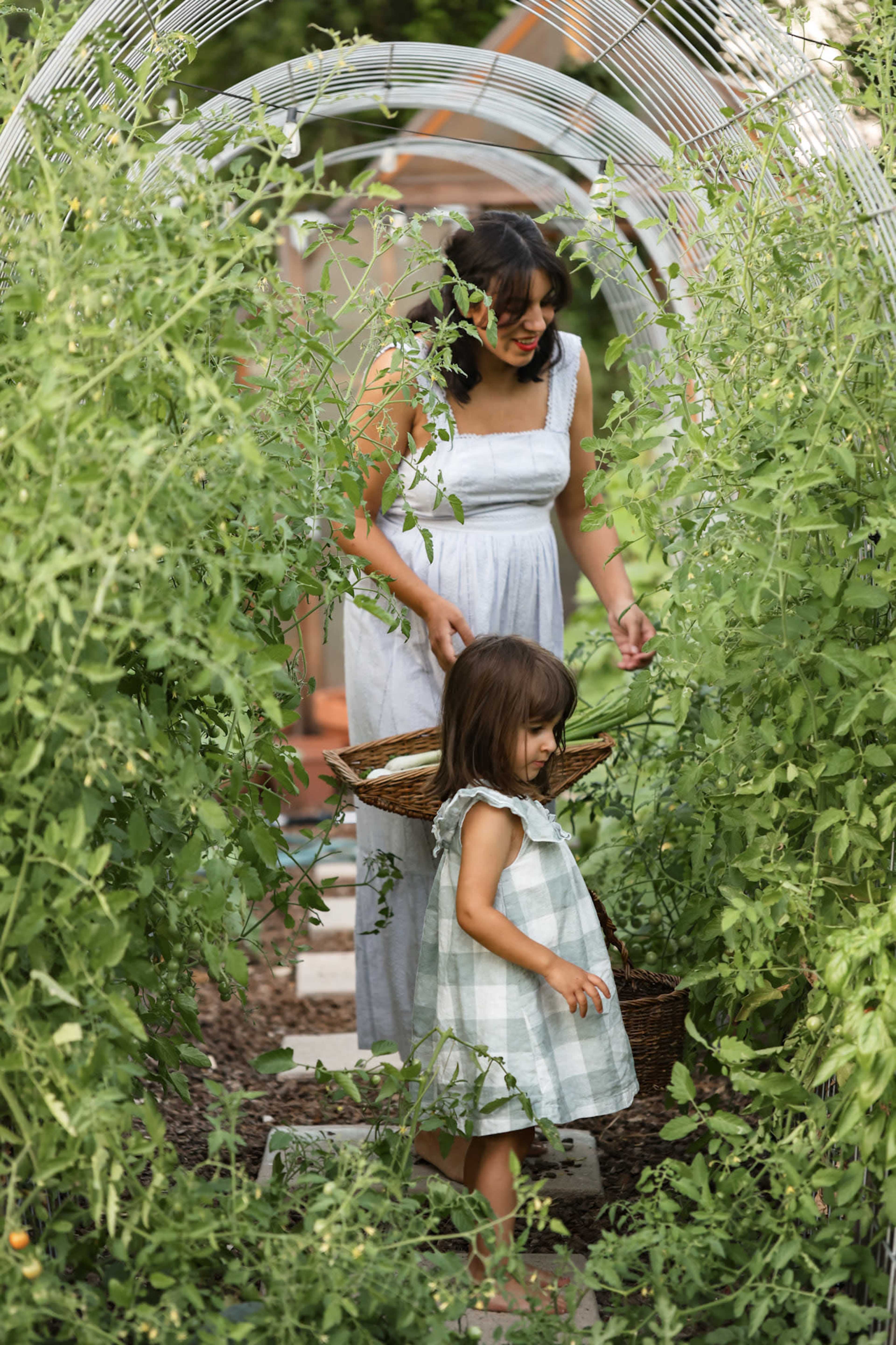 A woman and a girl walk through a garden, collecting vegetables in baskets surrounded by tall greenery.