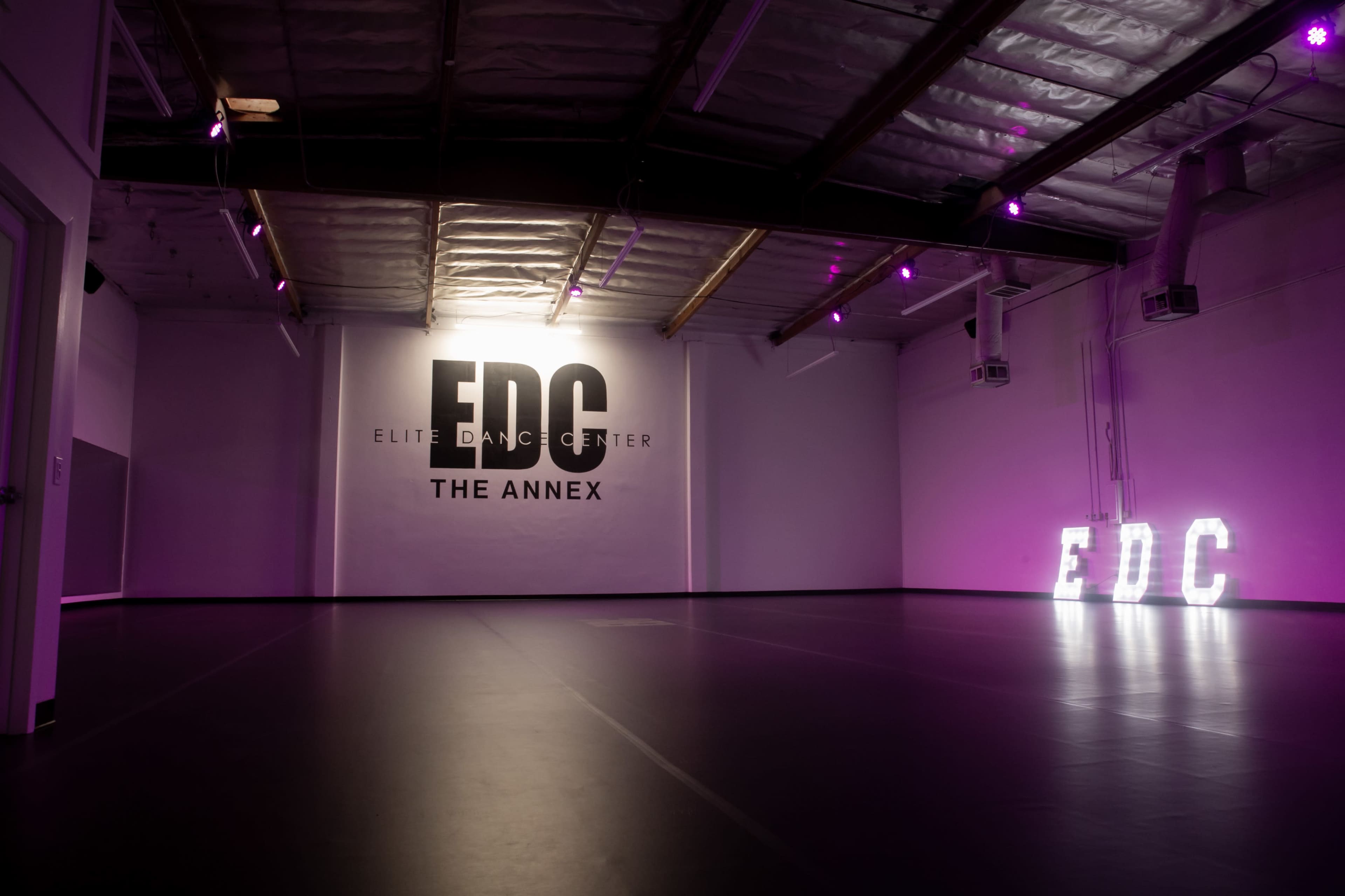 The interior of a dance studio featuring the words "EDC" and "THE ANNEX" on the wall, with illuminated letters spelling "EDC" on the floor and purple lighting.
