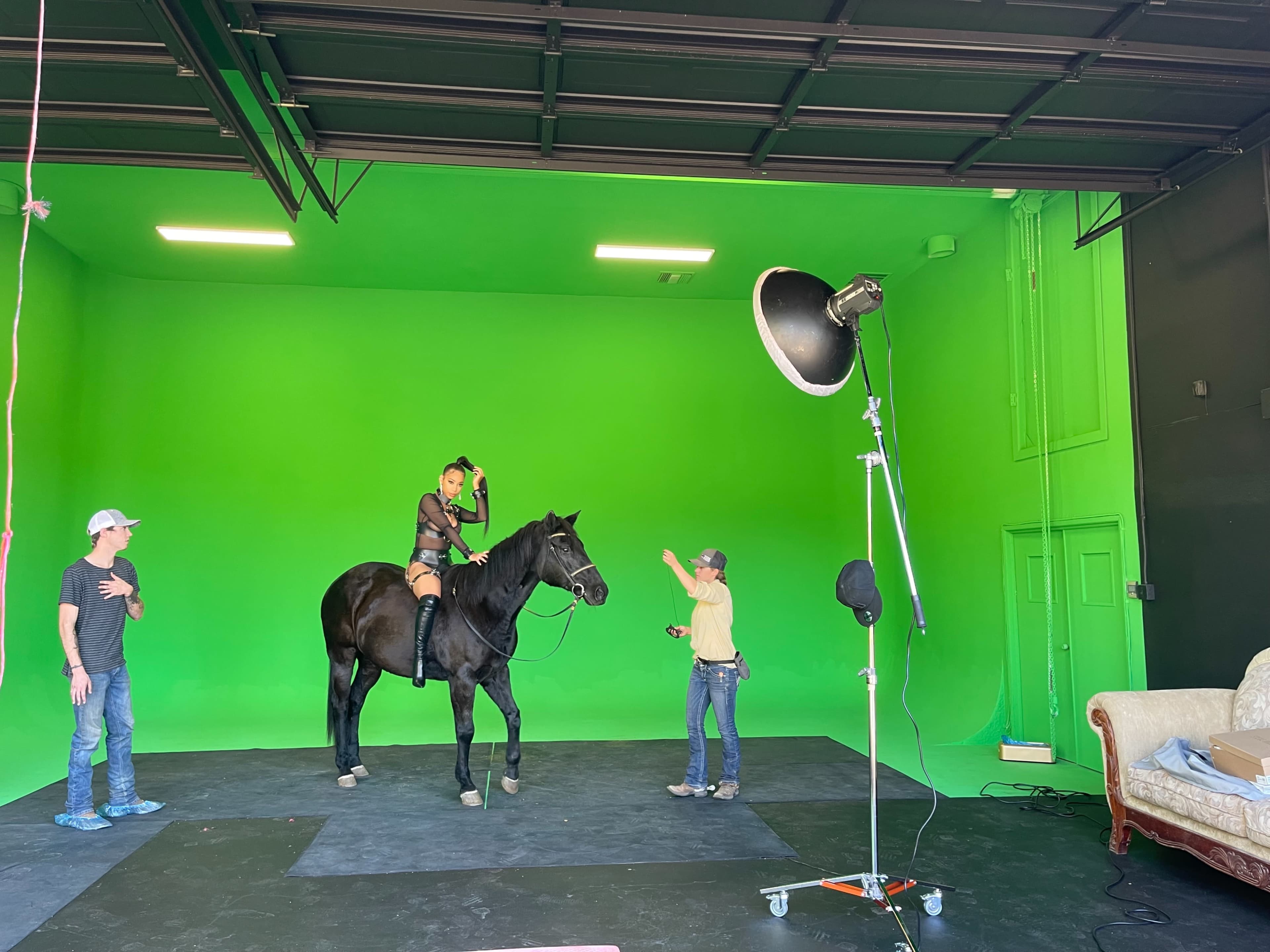 A person is riding a black horse inside a studio with a green screen background, while two crew members adjust equipment nearby.