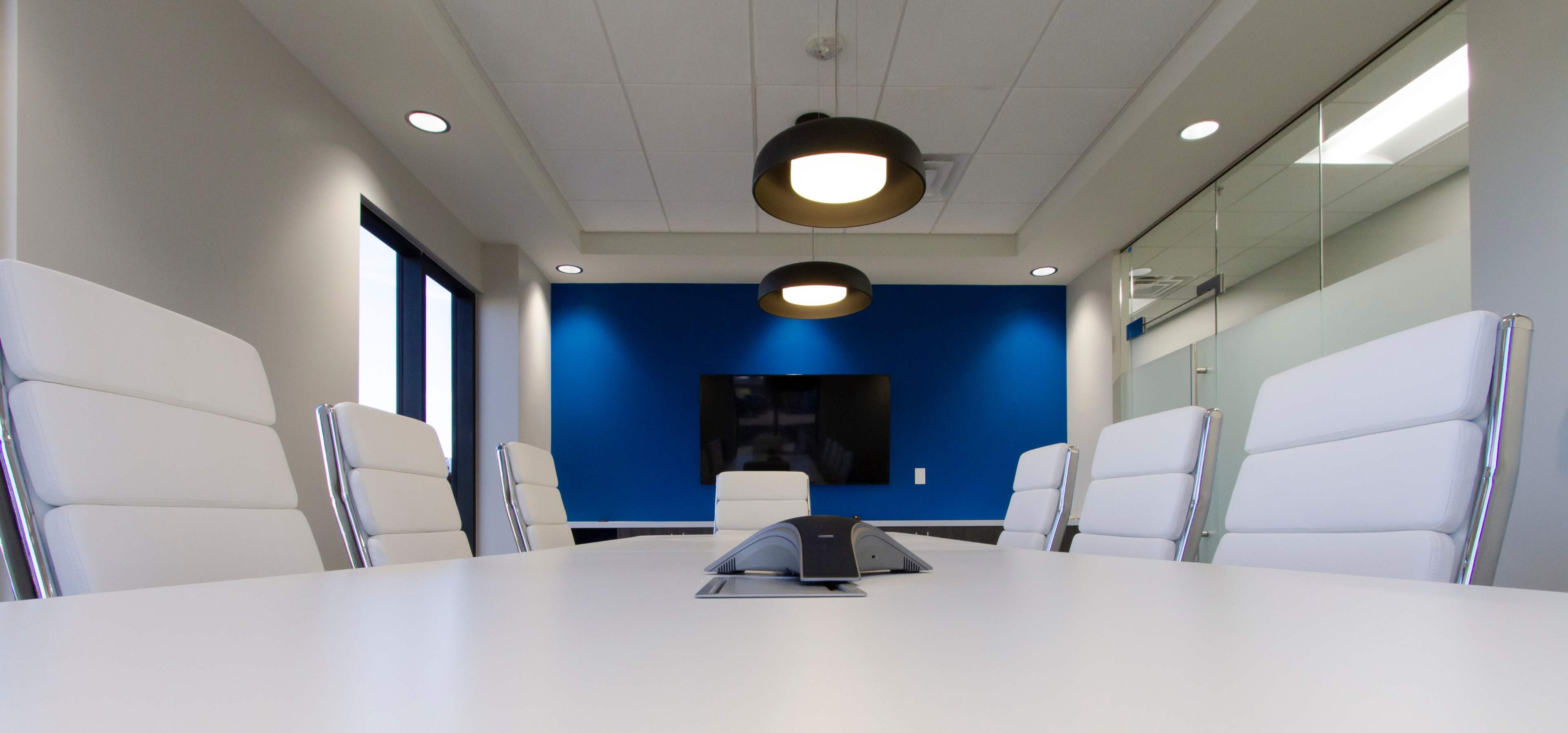 A modern conference room features a long white table surrounded by sleek white chairs, with a blue accent wall and a large screen at one end.
