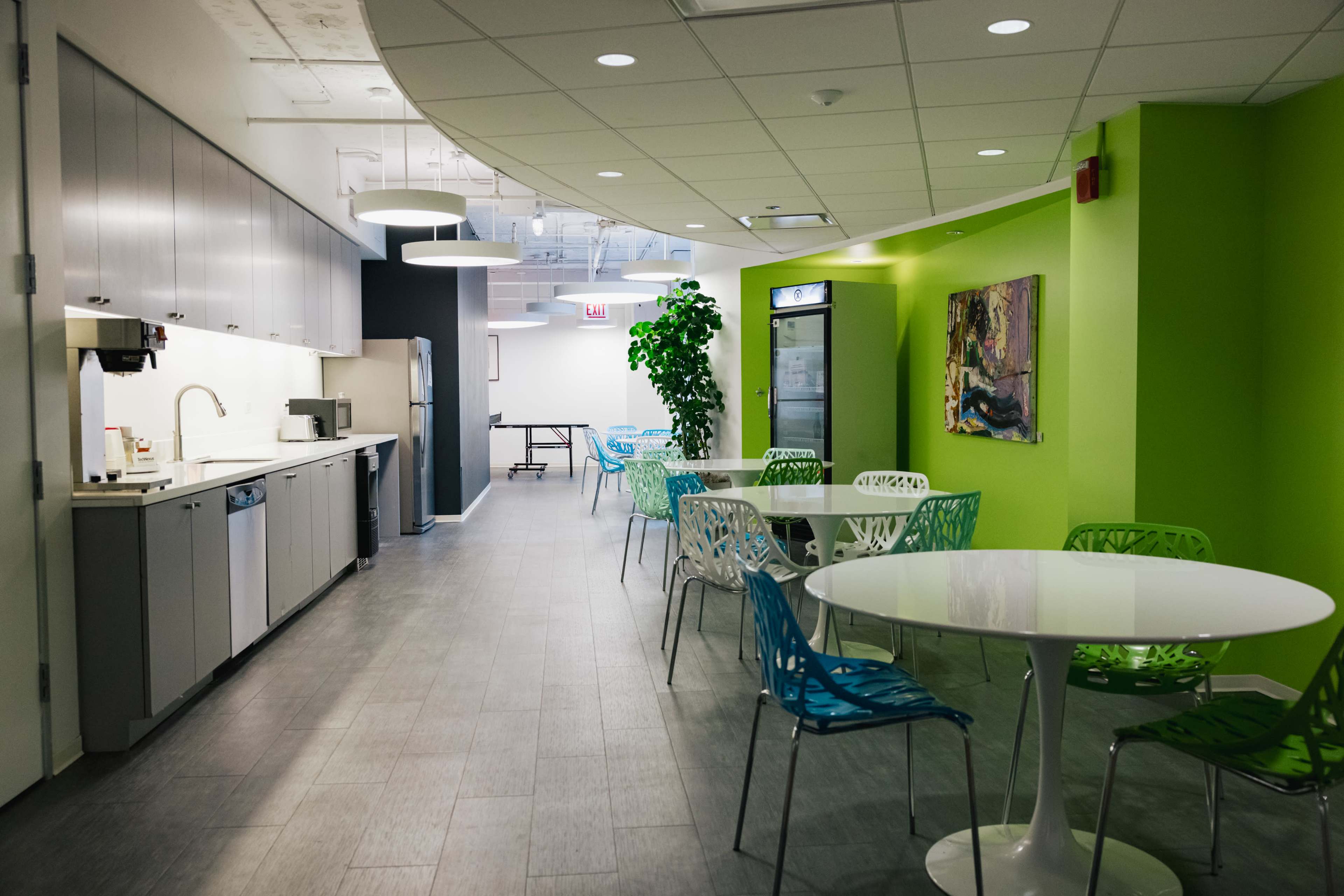 The image shows a modern break room with a kitchenette on one side, colorful chairs around white tables, and large green walls.