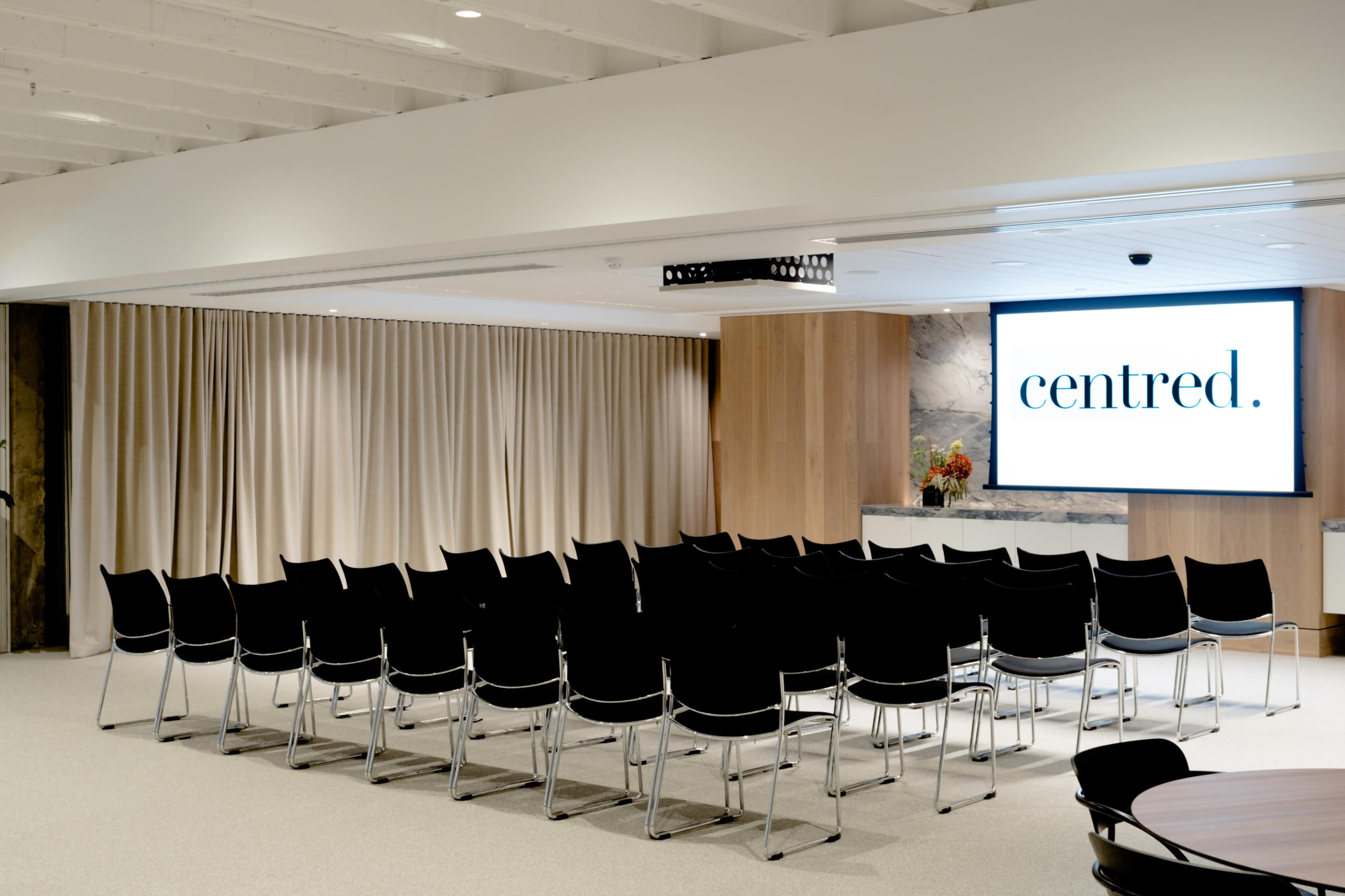 A modern meeting room with rows of black chairs facing a large screen displaying the word "centred," set against beige curtains and wooden paneling.
