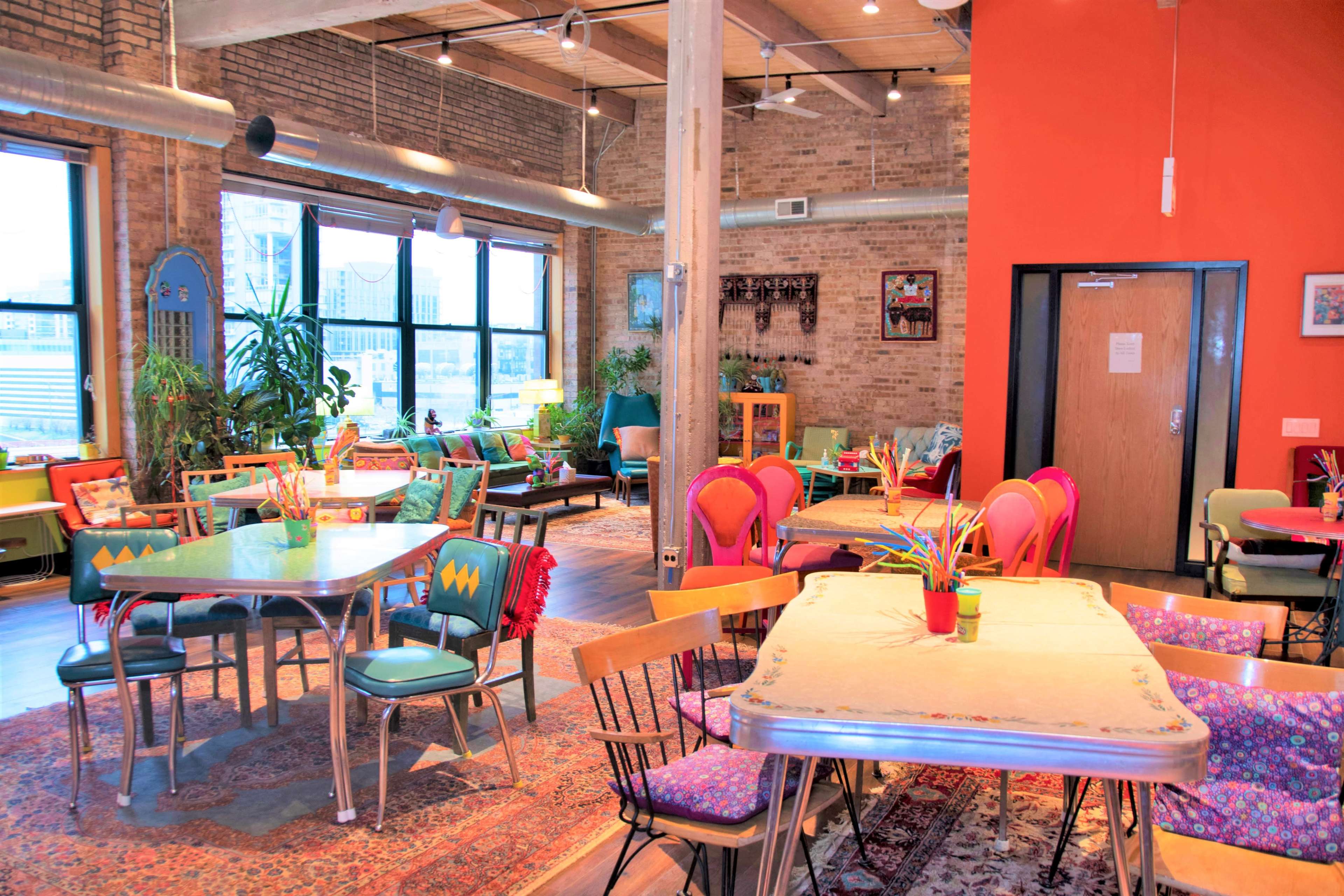 A brightly lit café with various seating arrangements, colorful chairs, and plants, set against a backdrop of exposed brick walls.