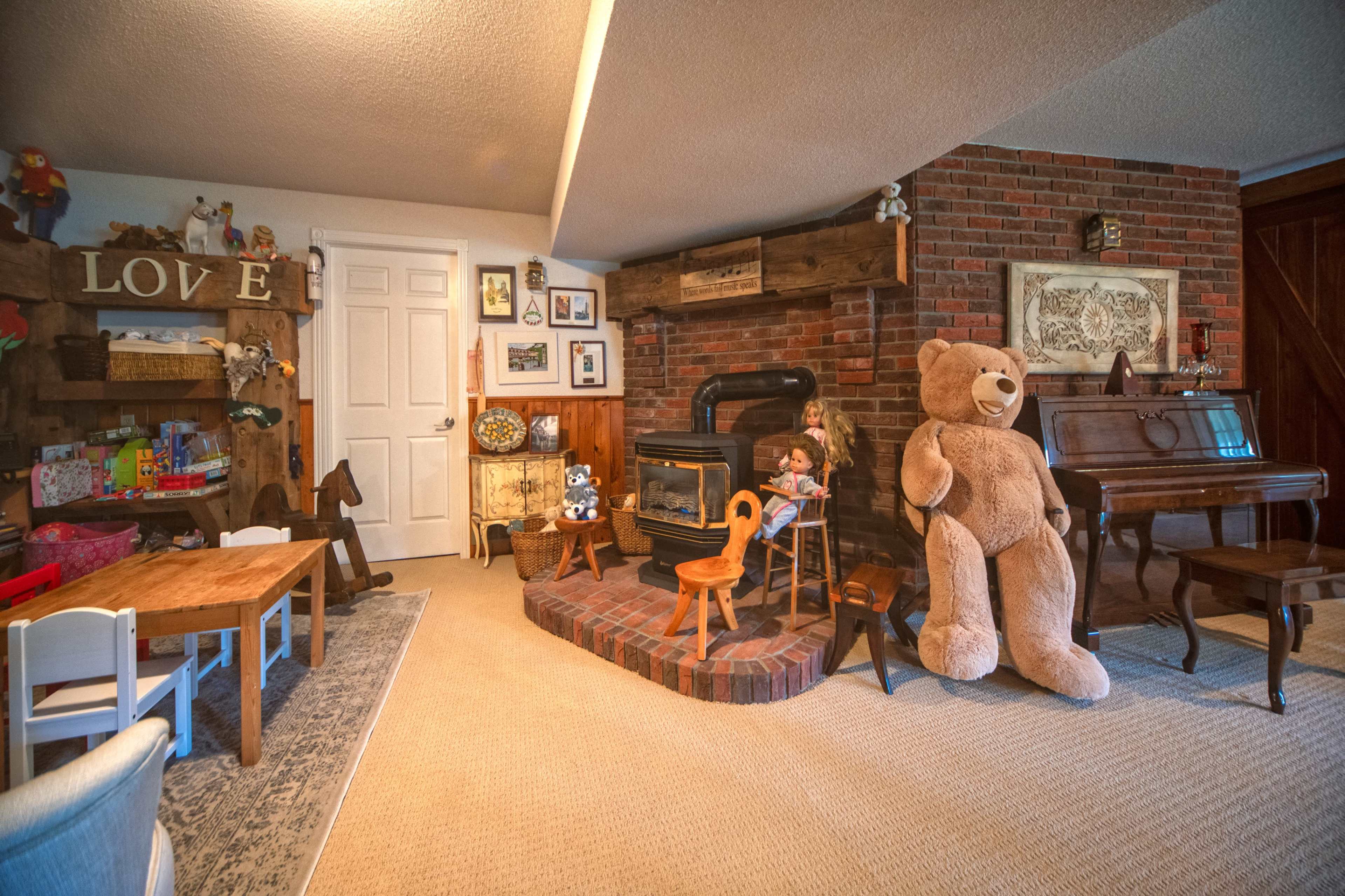 The image shows a cozy room with a brick fireplace, a large teddy bear seated nearby, a wooden table and chairs, and various decorations on the walls and shelves.