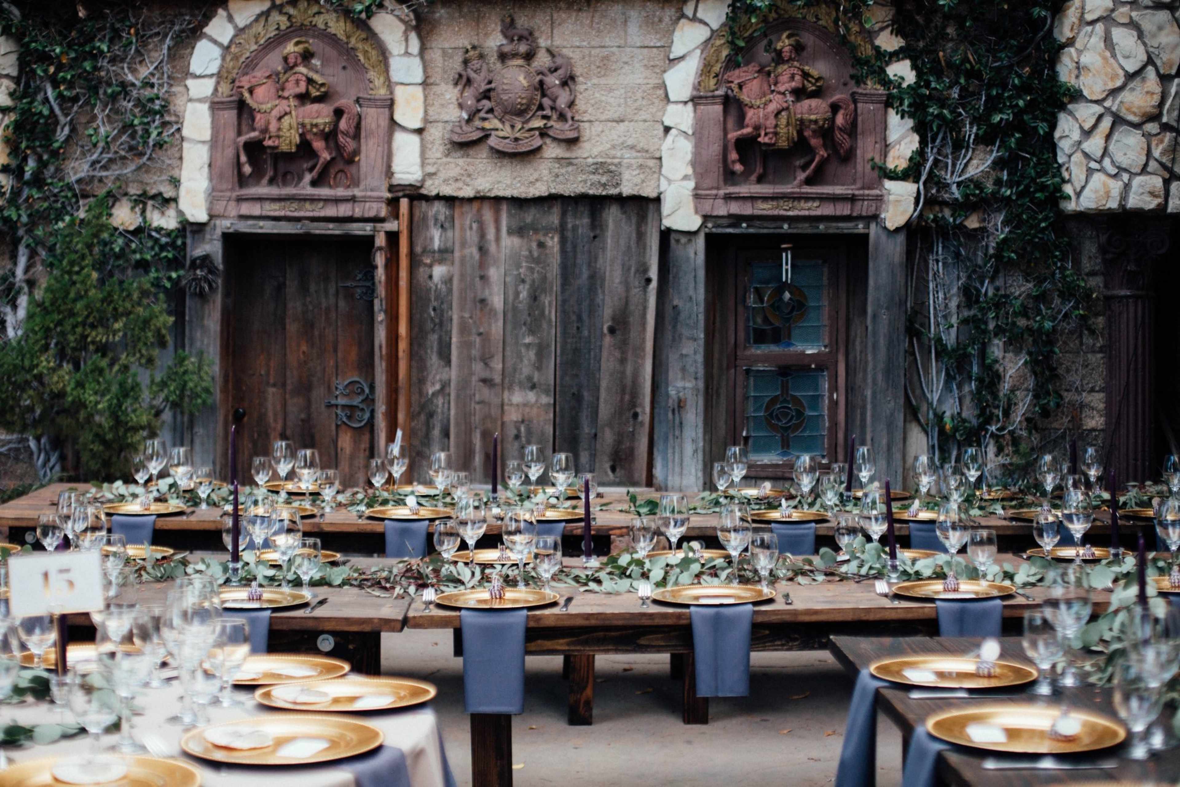 A rustic dining area with long wooden tables set for a formal meal, featuring elegant tableware, foliage decorations, and stone walls adorned with ornate carvings.