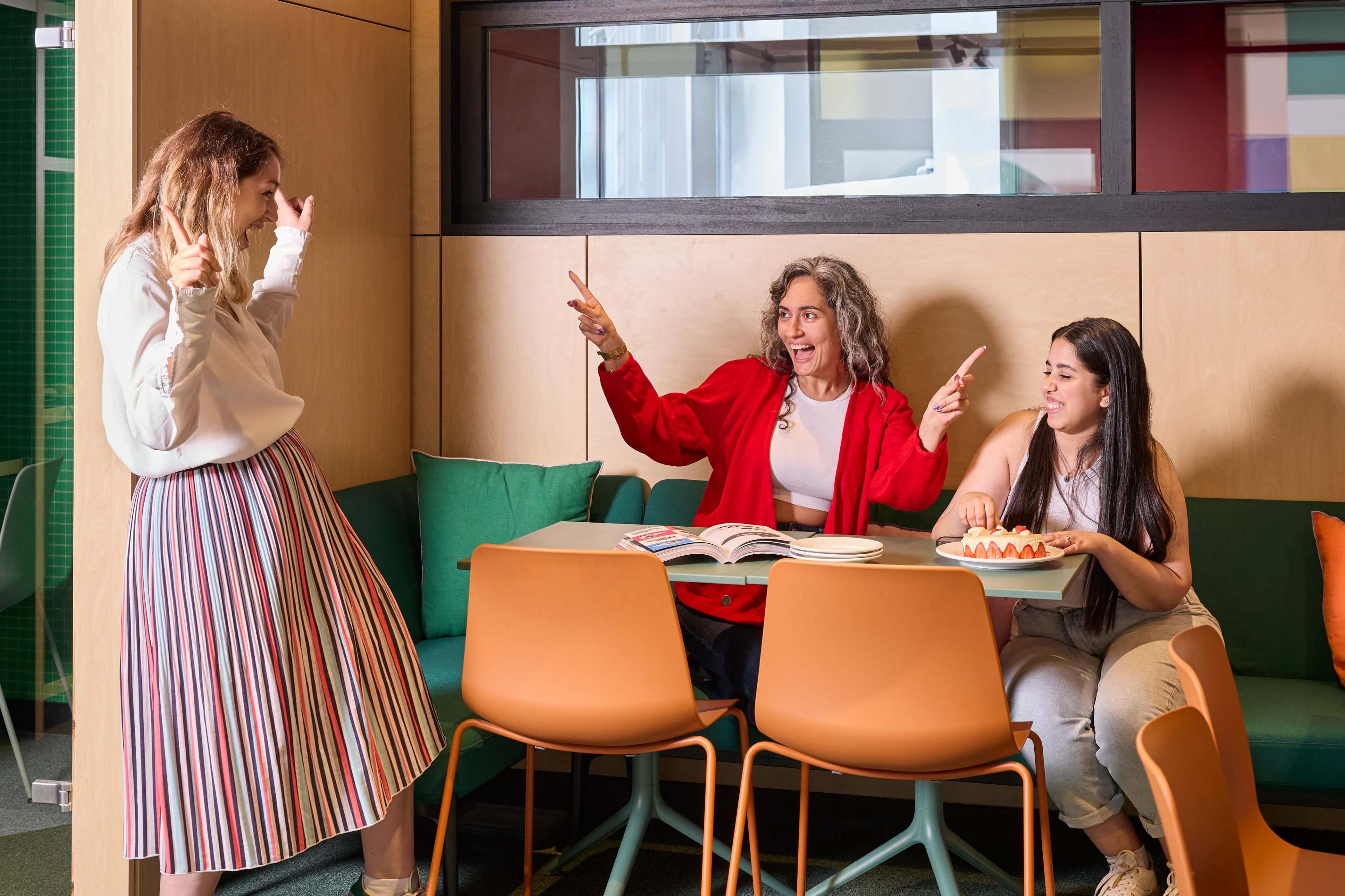 Three women are engaged in a lively conversation at a table in a brightly colored, modern workspace.