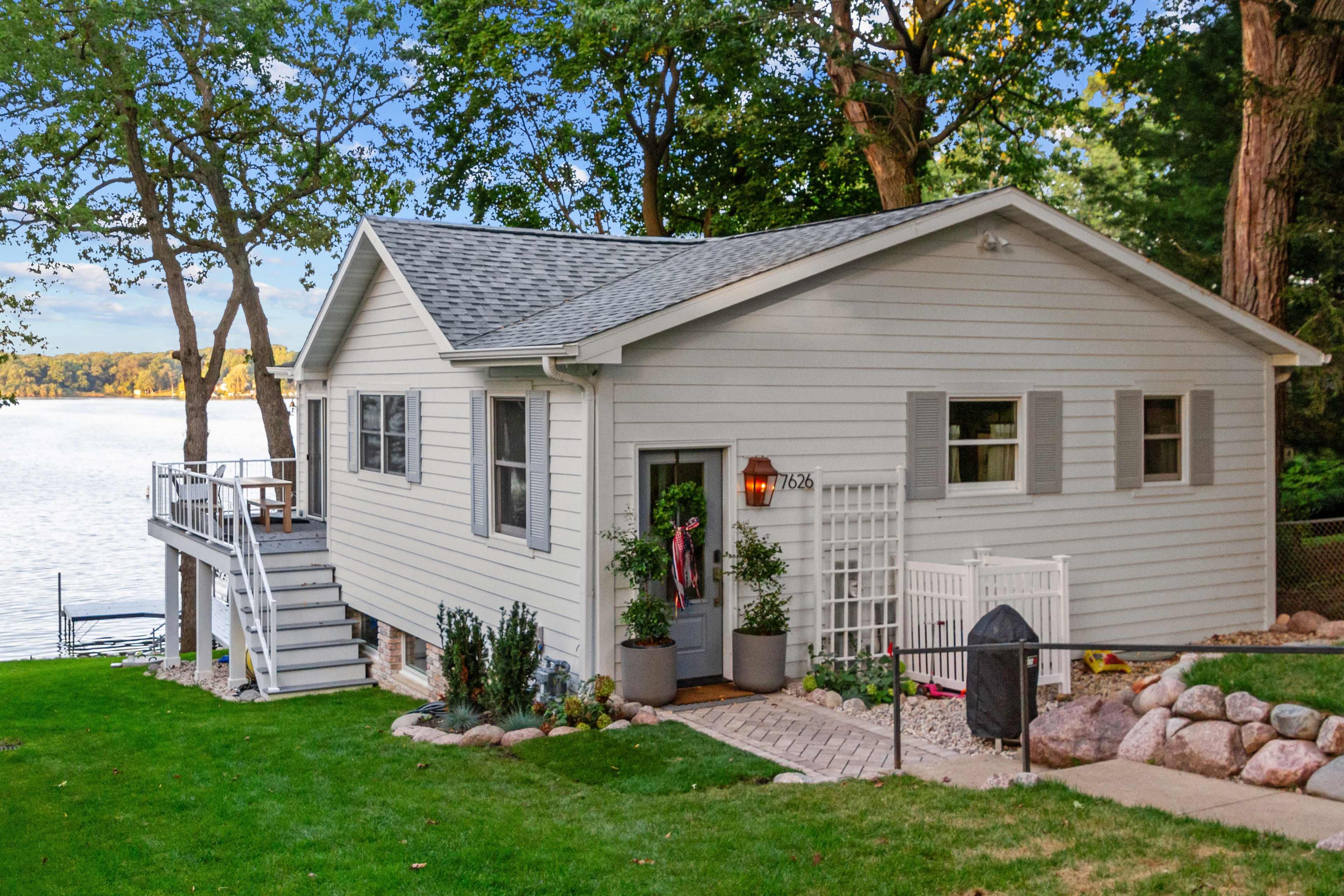 A light gray house with a deck overlooks a calm lake, surrounded by trees and a landscaped garden.