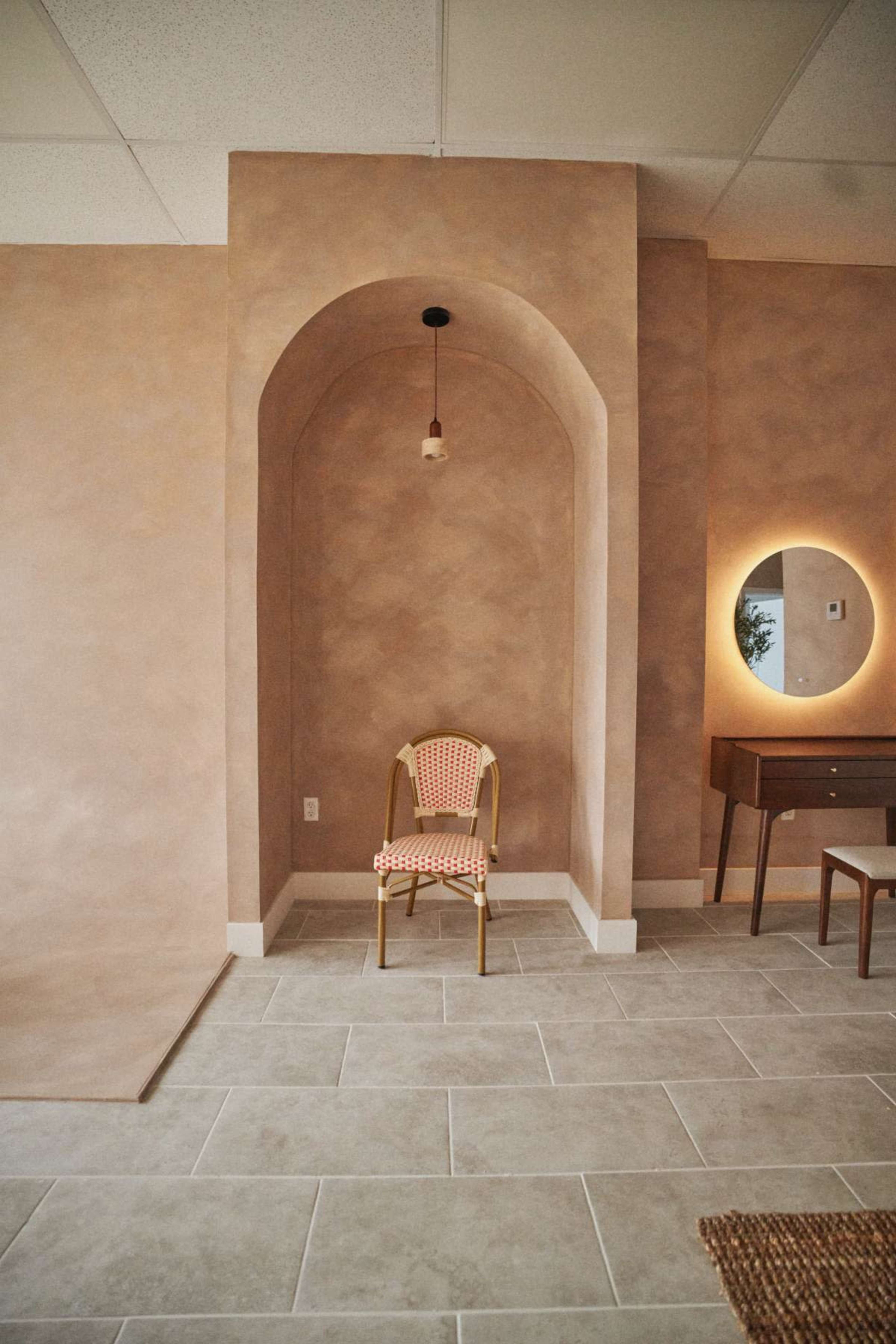 The image shows a minimalist interior space with a textured beige wall, an arched alcove containing a patterned chair, and a circular light fixture near a wooden vanity.