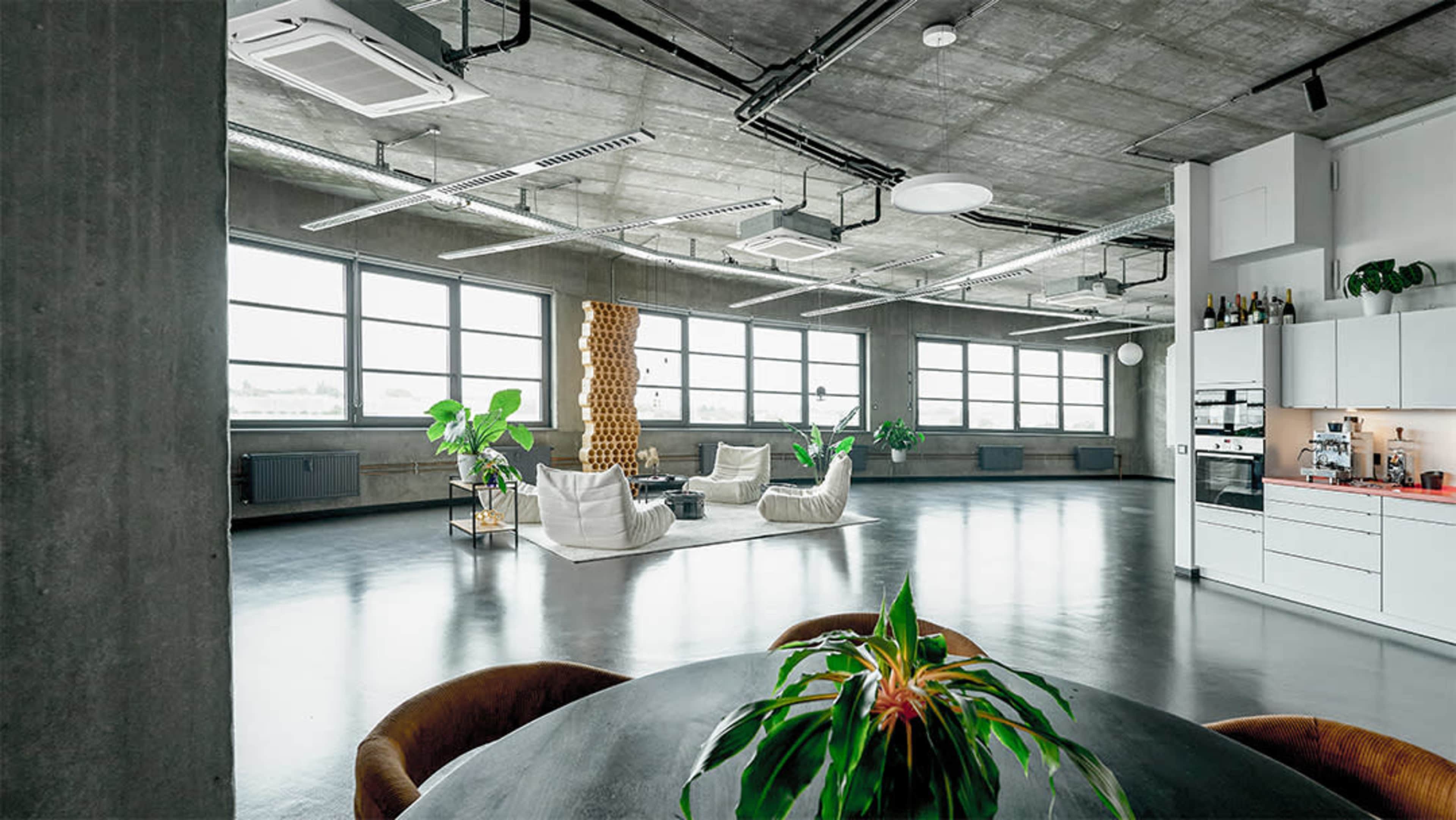 The image shows a spacious, industrial-style room with large windows, concrete walls, and minimalist furniture, including two white chairs and a round dining table with a plant centerpiece.