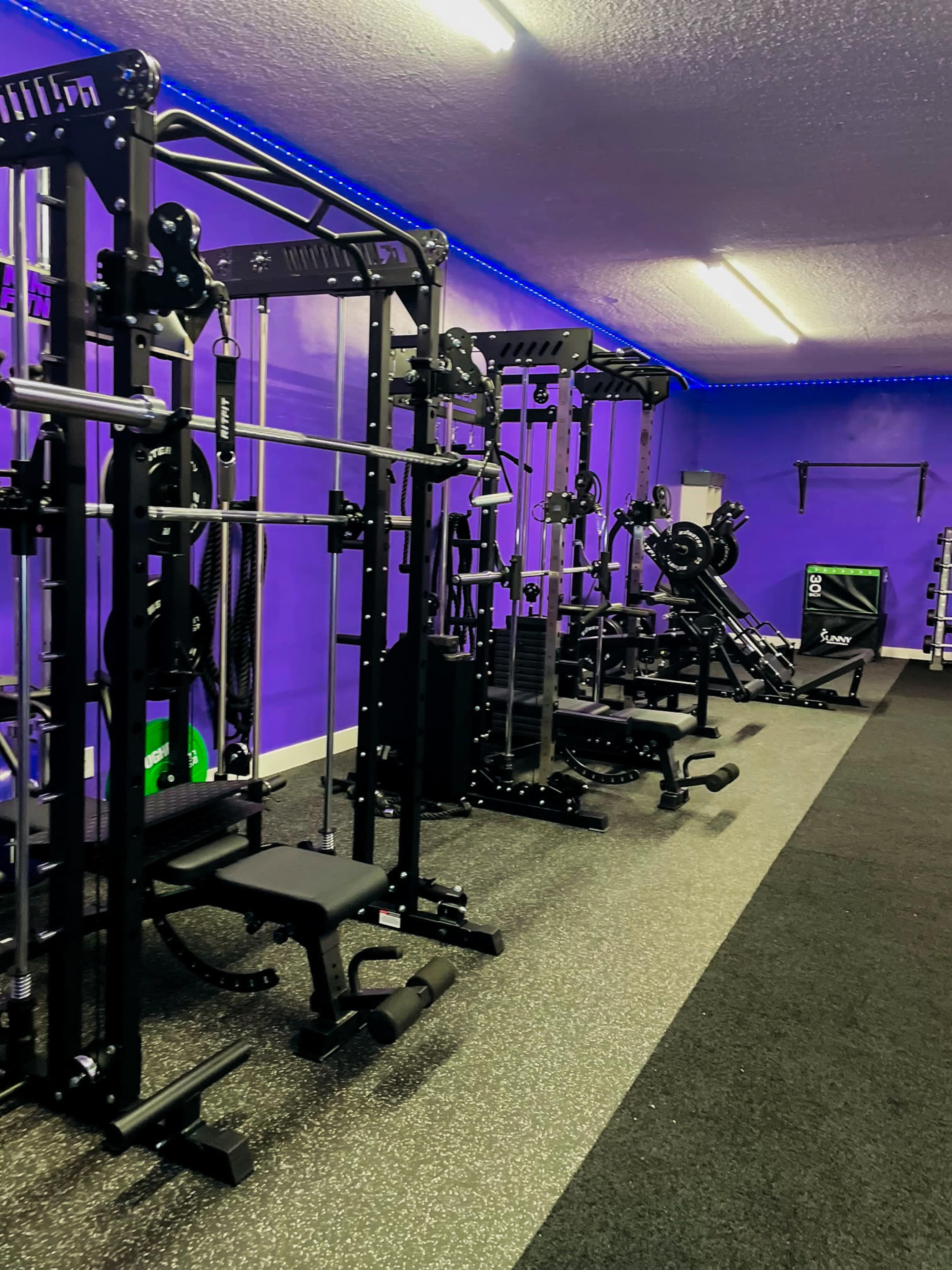 The image shows a modern gym interior with various weightlifting and exercise machines, illuminated by blue LED lighting against a purple wall.