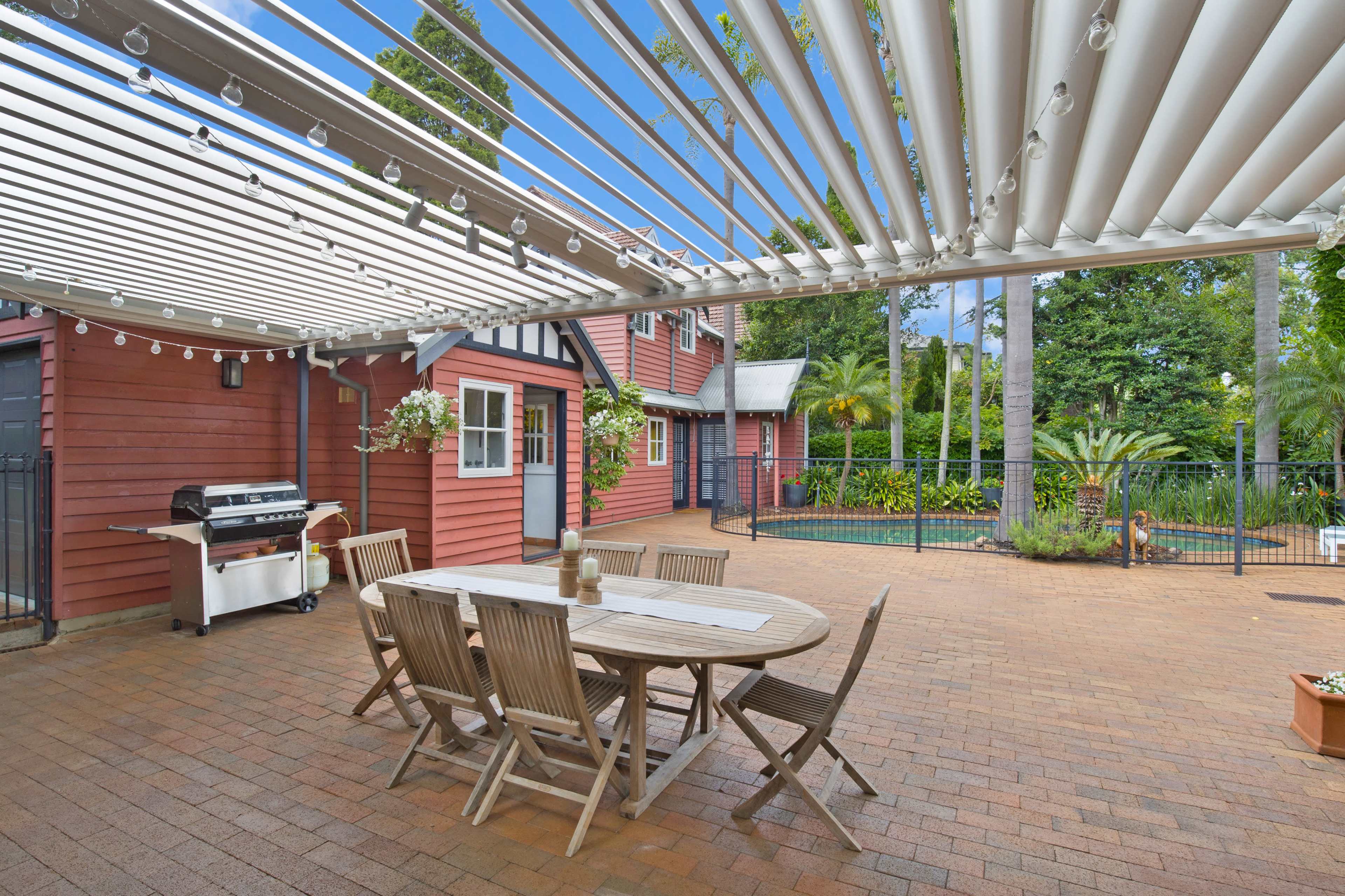 An outdoor patio with a wooden dining table and chairs is covered by a louvered pergola, next to a swimming pool and surrounded by landscaped greenery.