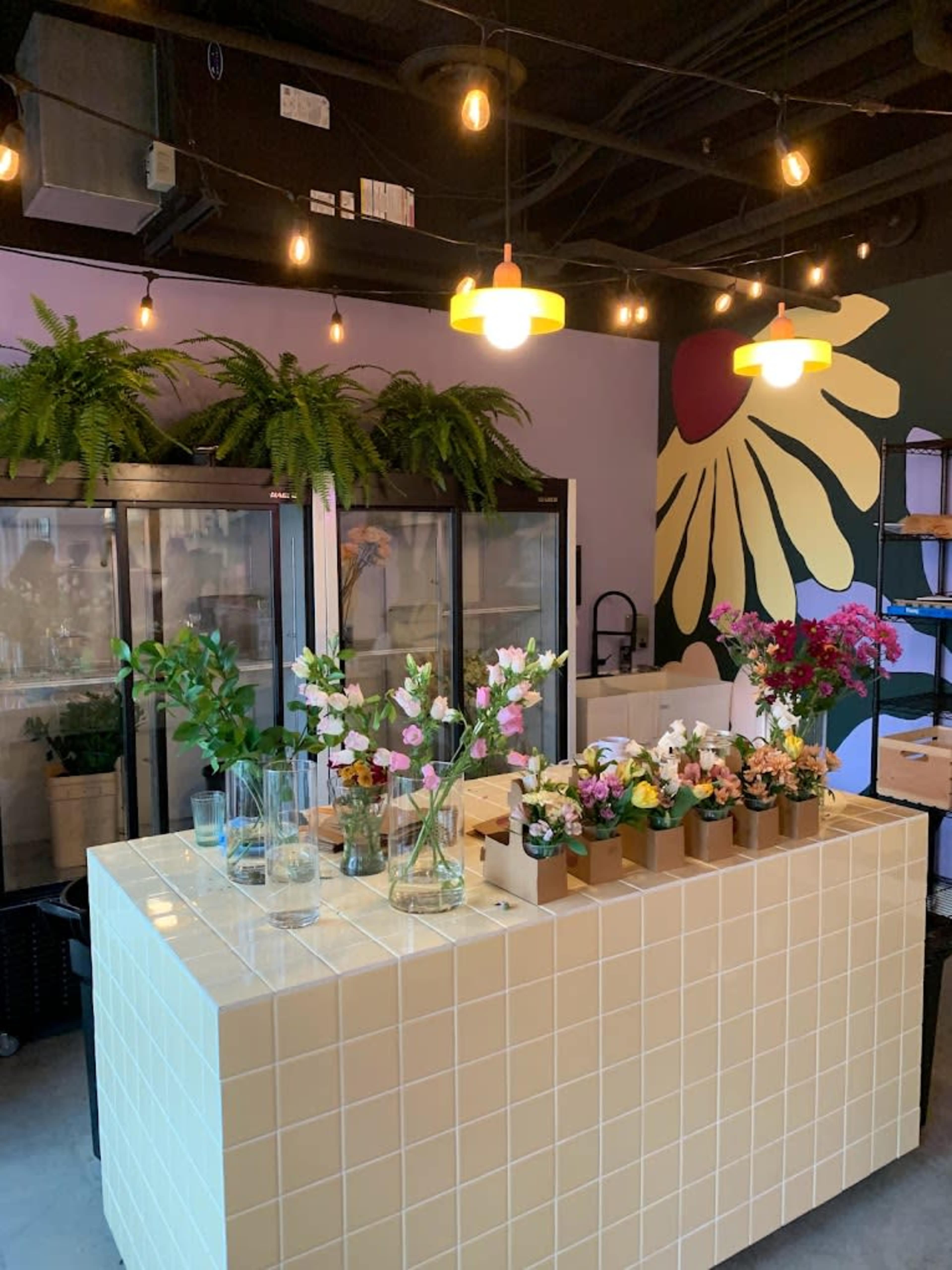 A floral shop features a tiled counter adorned with various potted flowers and plants, surrounded by glass display cases and hanging lights.