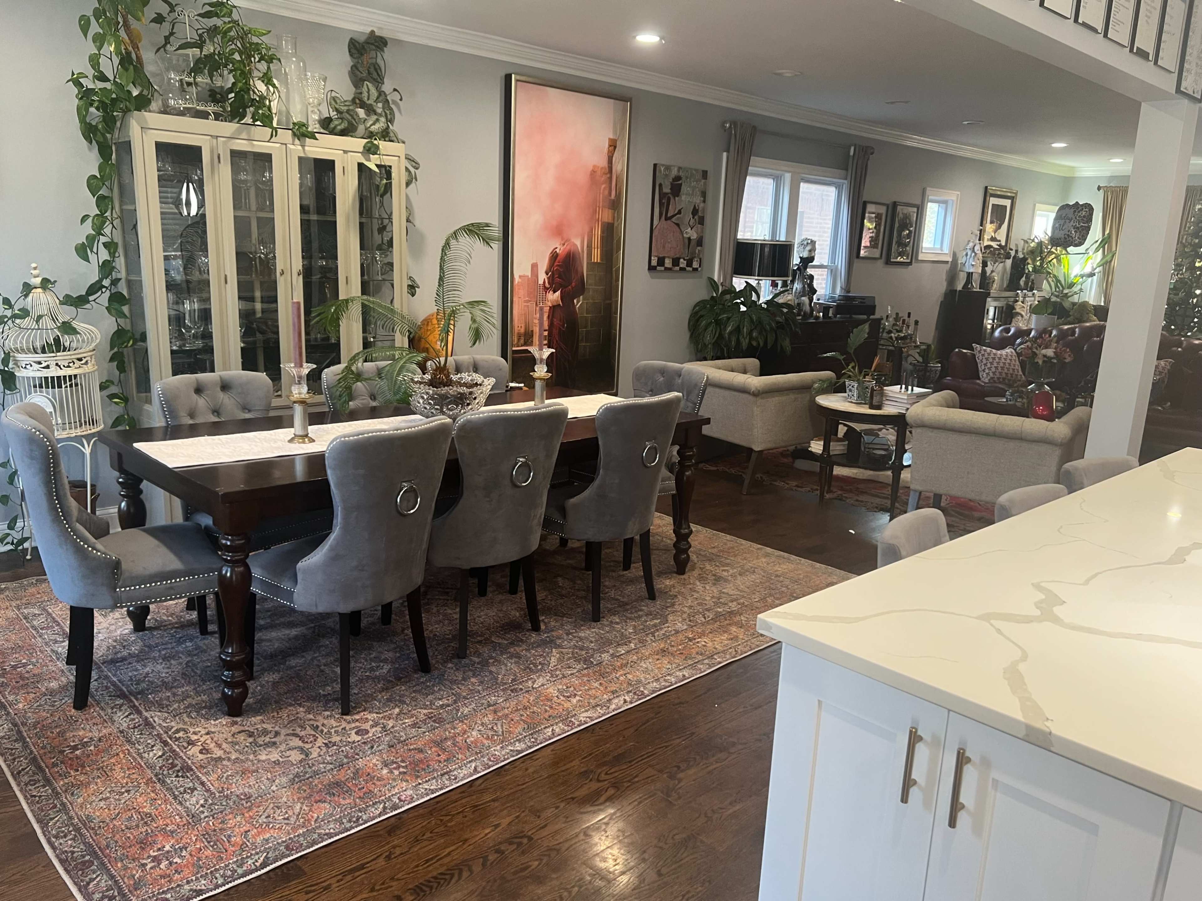 The image shows a dining area with a large wooden table surrounded by gray upholstered chairs, complemented by a decorative rug and various plants, in a well-lit, open-concept living space.