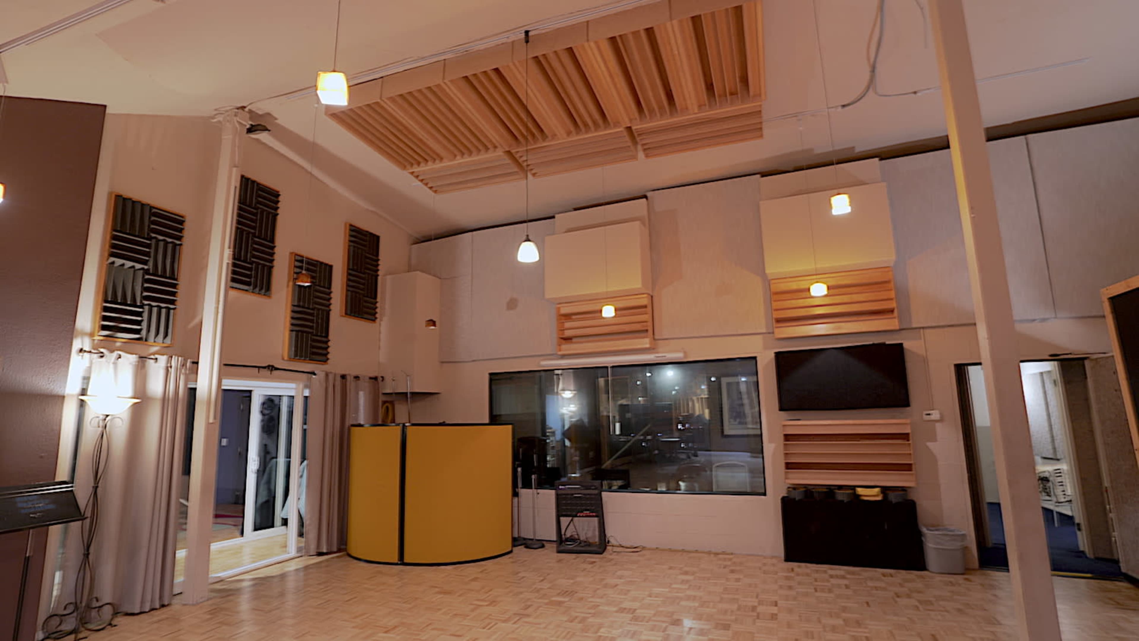A sound studio with acoustic panels, a control booth with a glass window, and a spacious wooden floor.