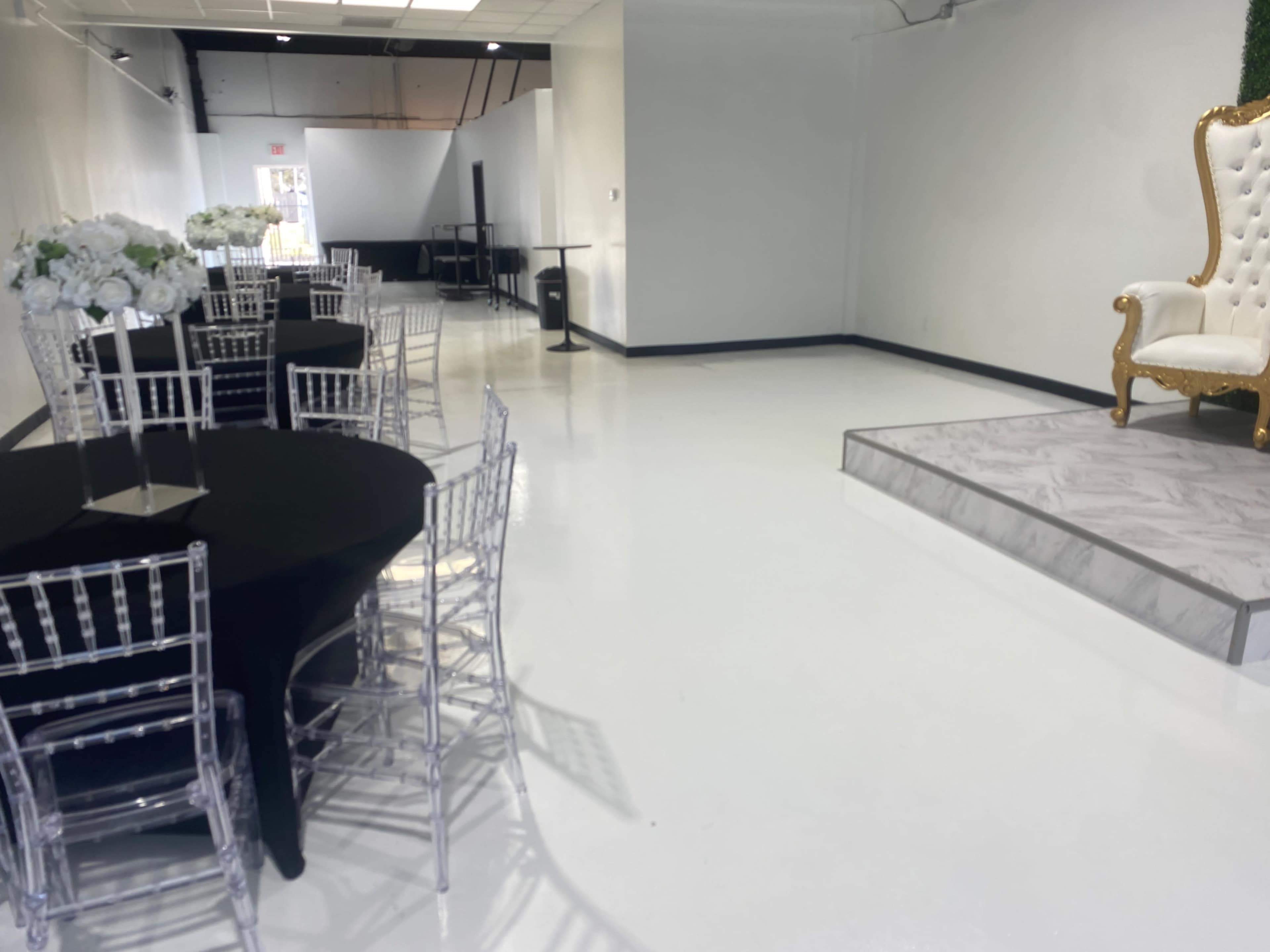 The image shows a spacious white-walled event room with black and clear chairs arranged around circular tables, and a raised platform with a decorative throne on one side.