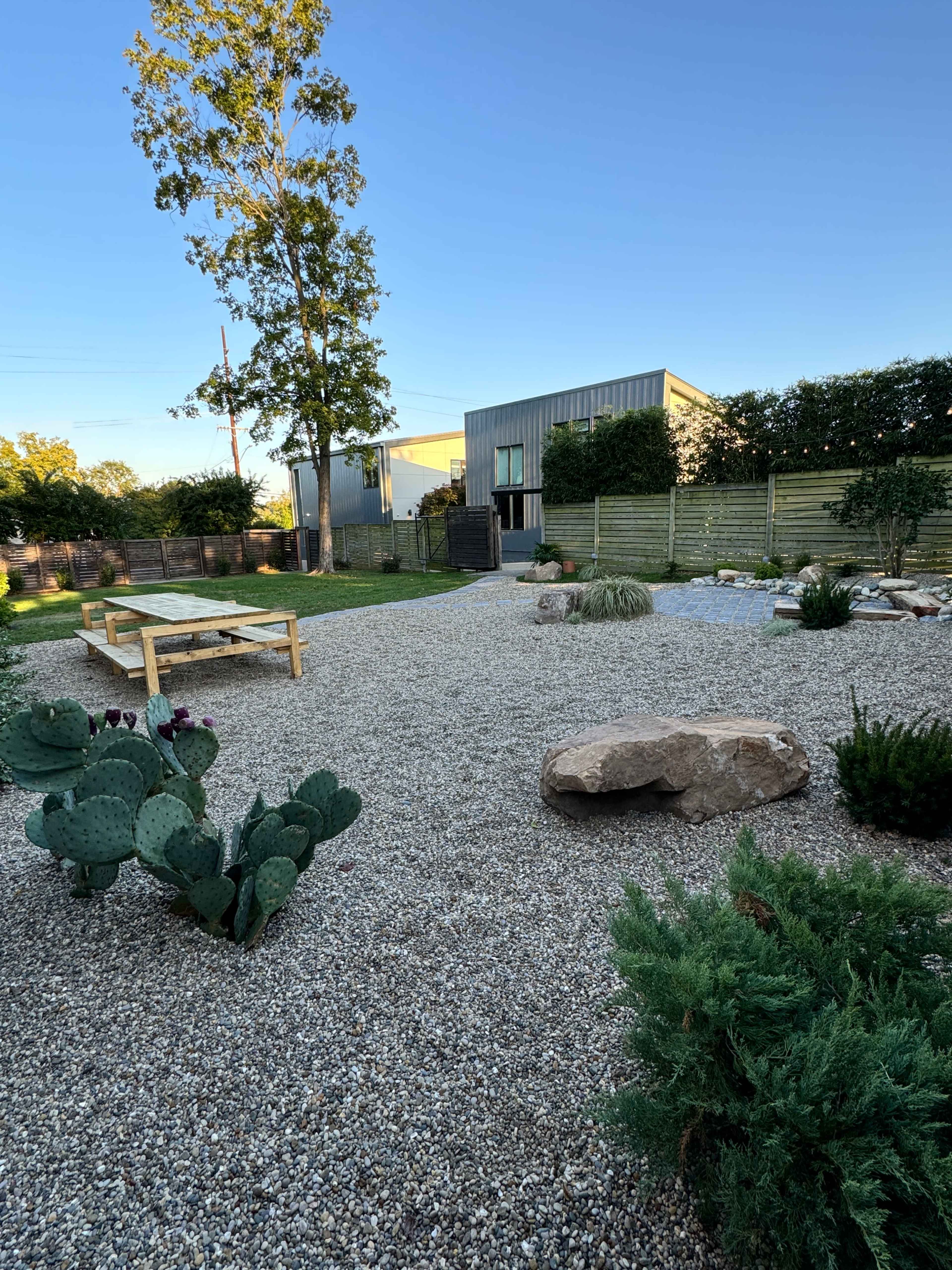 The image shows a landscaped garden featuring gravel, various plants including cacti, a wooden table, and a modern building in the background.
