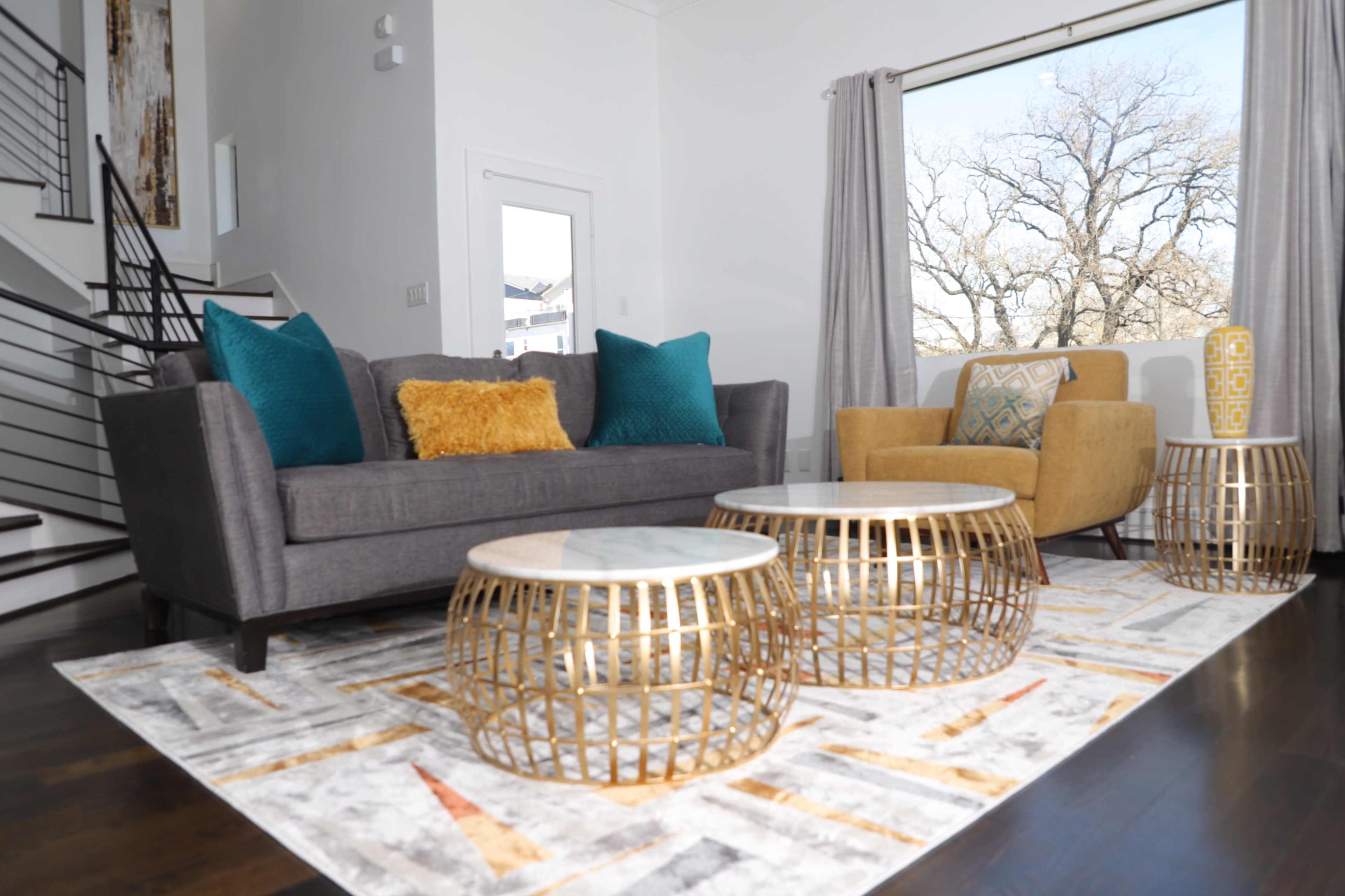A modern living room with a gray sofa, an armchair, two decorative gold tables, and a patterned rug, all set against a large window.
