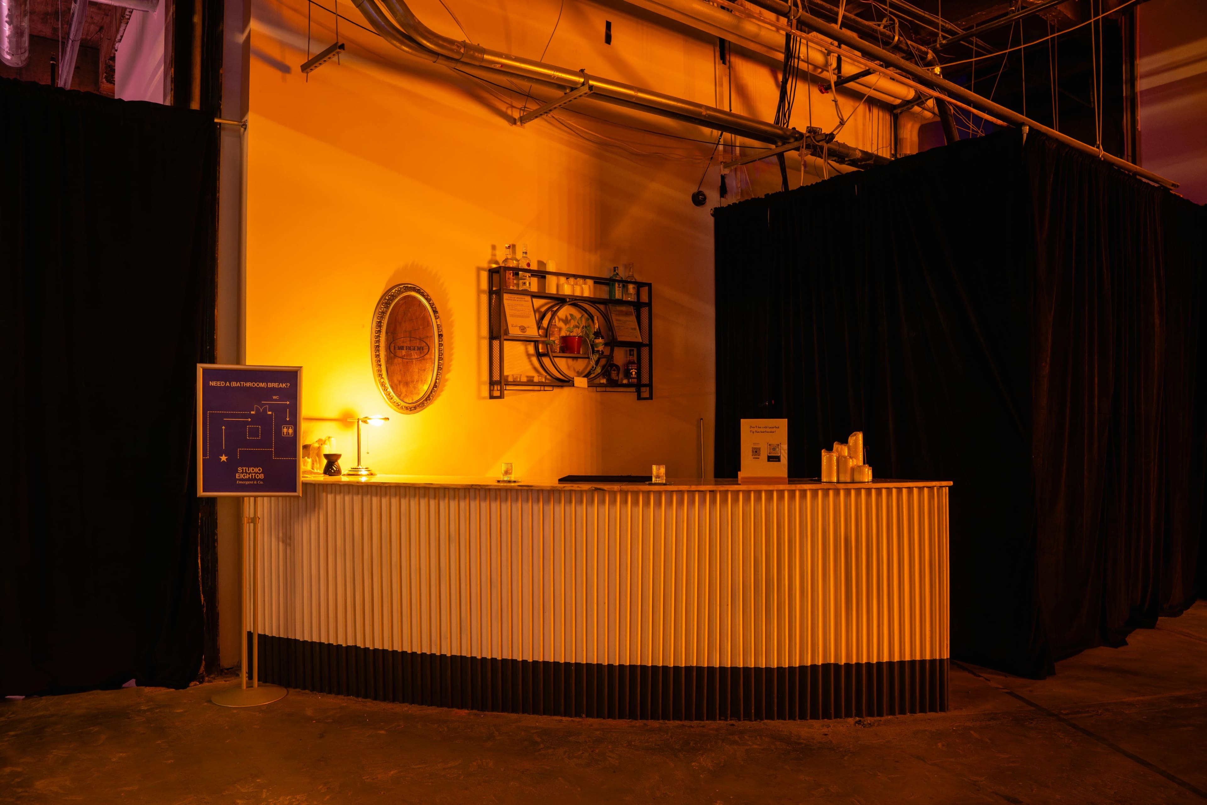 A reception desk is illuminated with warm lighting, featuring a sign nearby and surrounded by black curtains in a spacious area.
