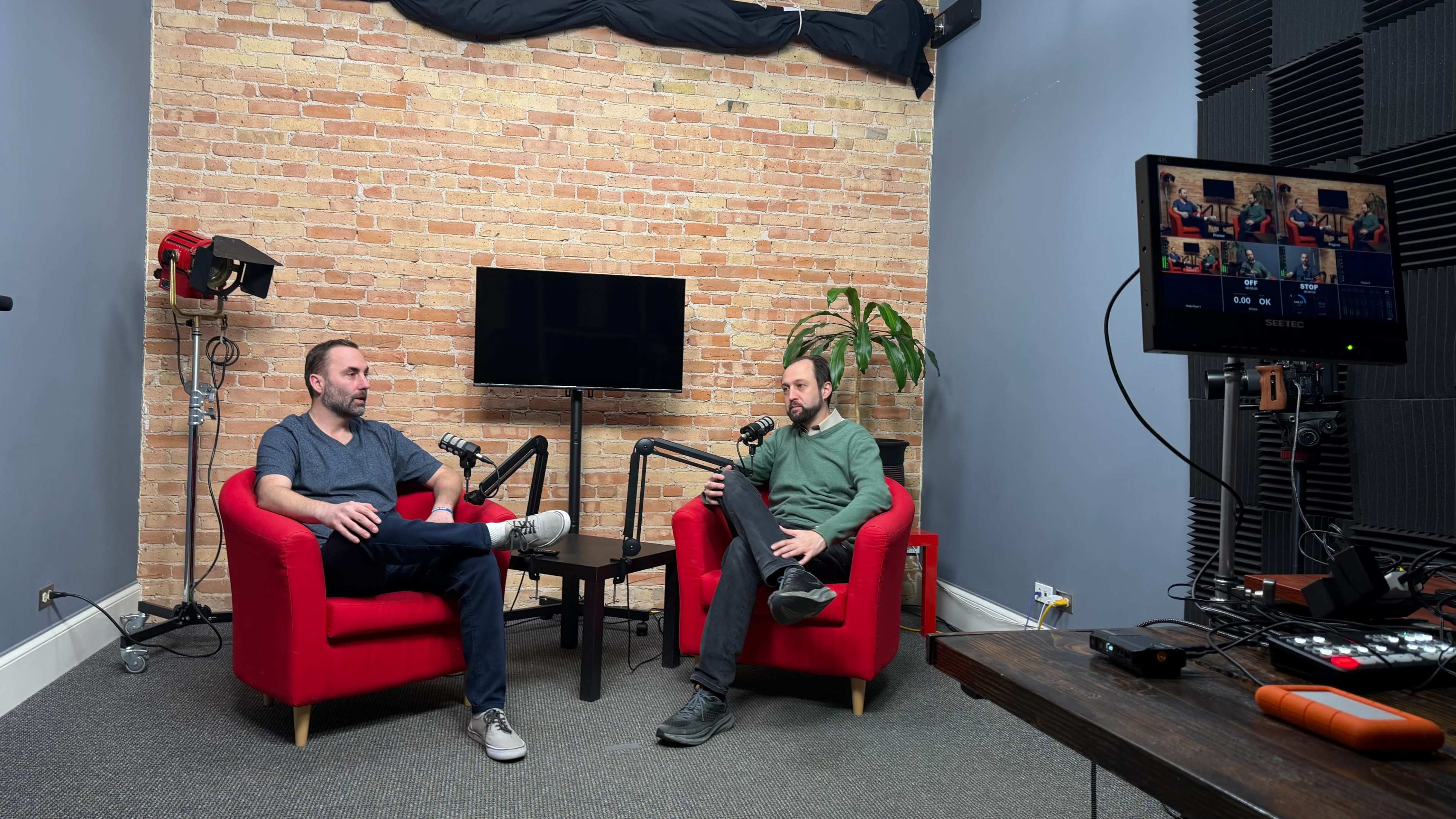 Two men are seated in red armchairs in front of a brick wall, with a television screen and a potted plant visible in the background.