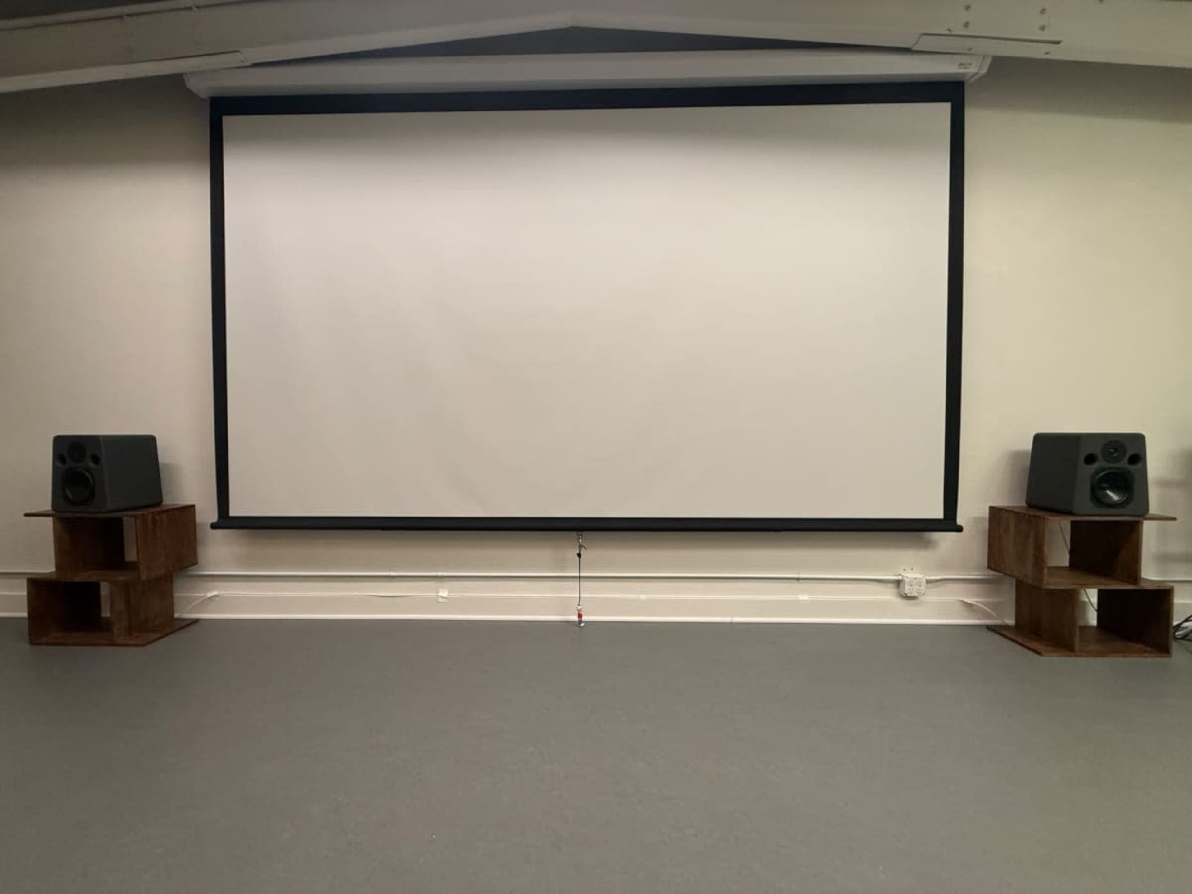 The image shows a blank projection screen in a room with two loudspeakers on wooden stands positioned on either side.