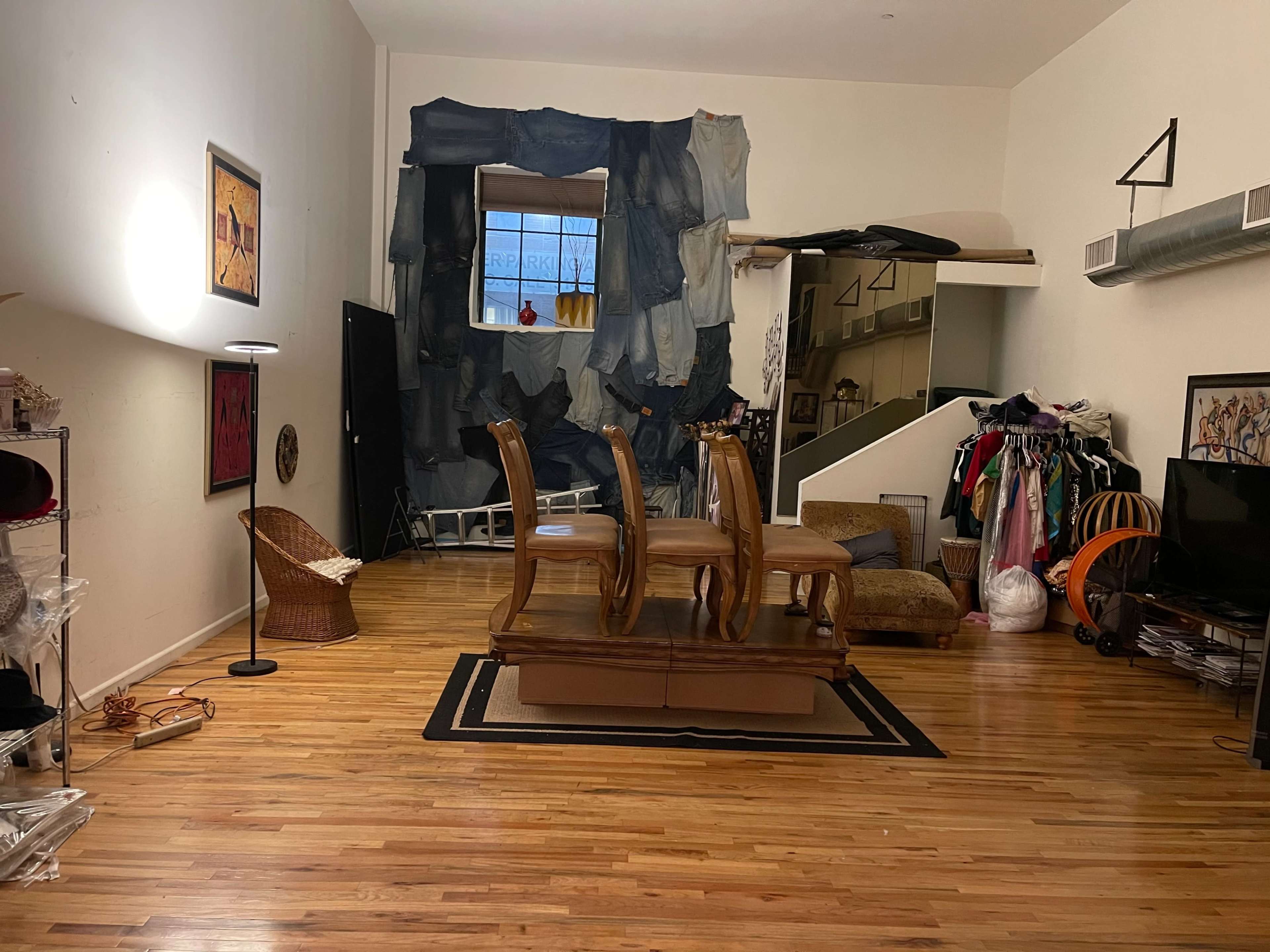 A spacious room with wooden flooring, featuring a table with four chairs at its center, a large piece of fabric art on the wall, and various furniture and items scattered around, including a floor lamp and a decorative chair.