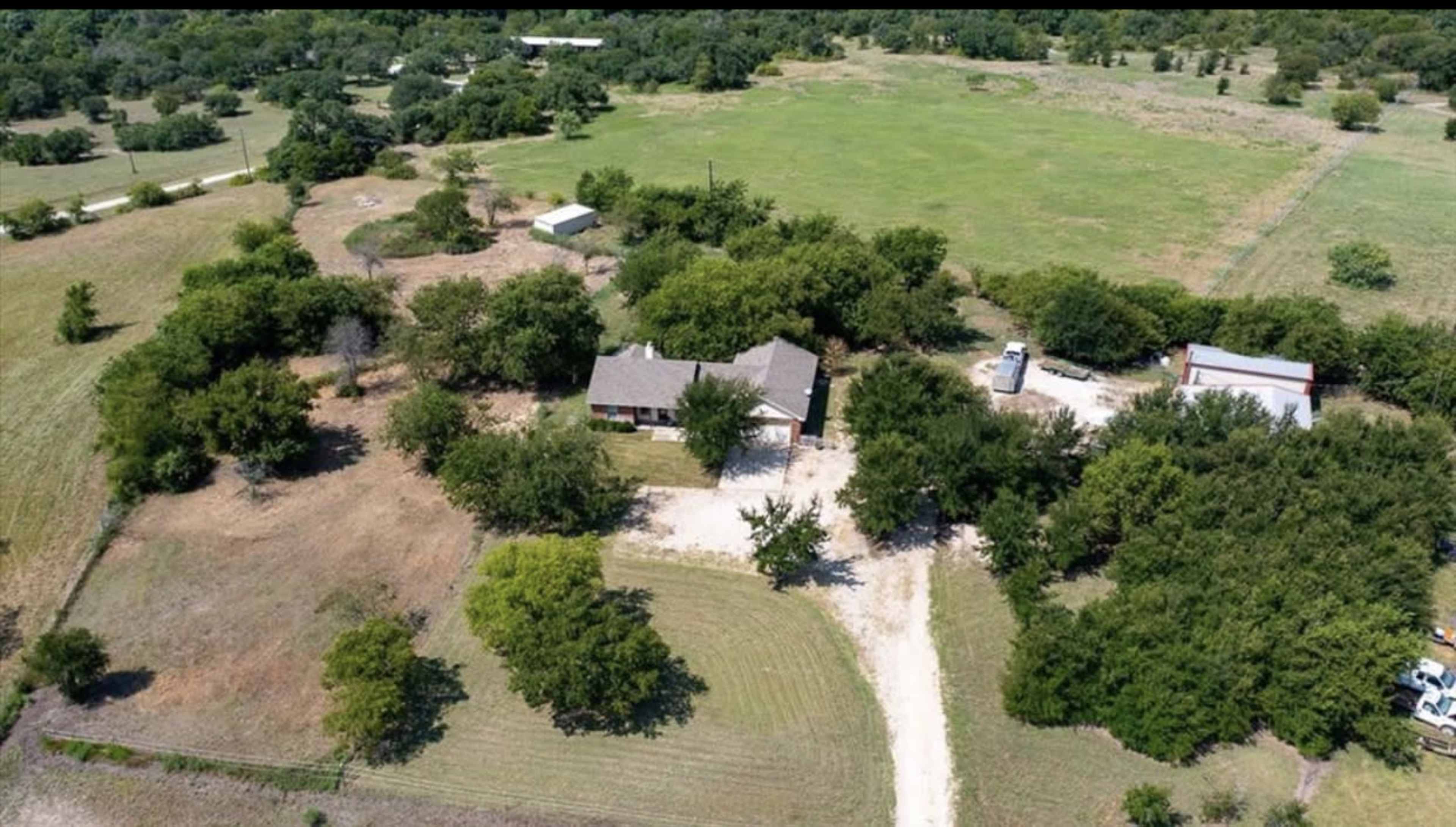 The image shows an aerial view of a rural property featuring a house surrounded by trees, with a dirt driveway and open fields nearby.