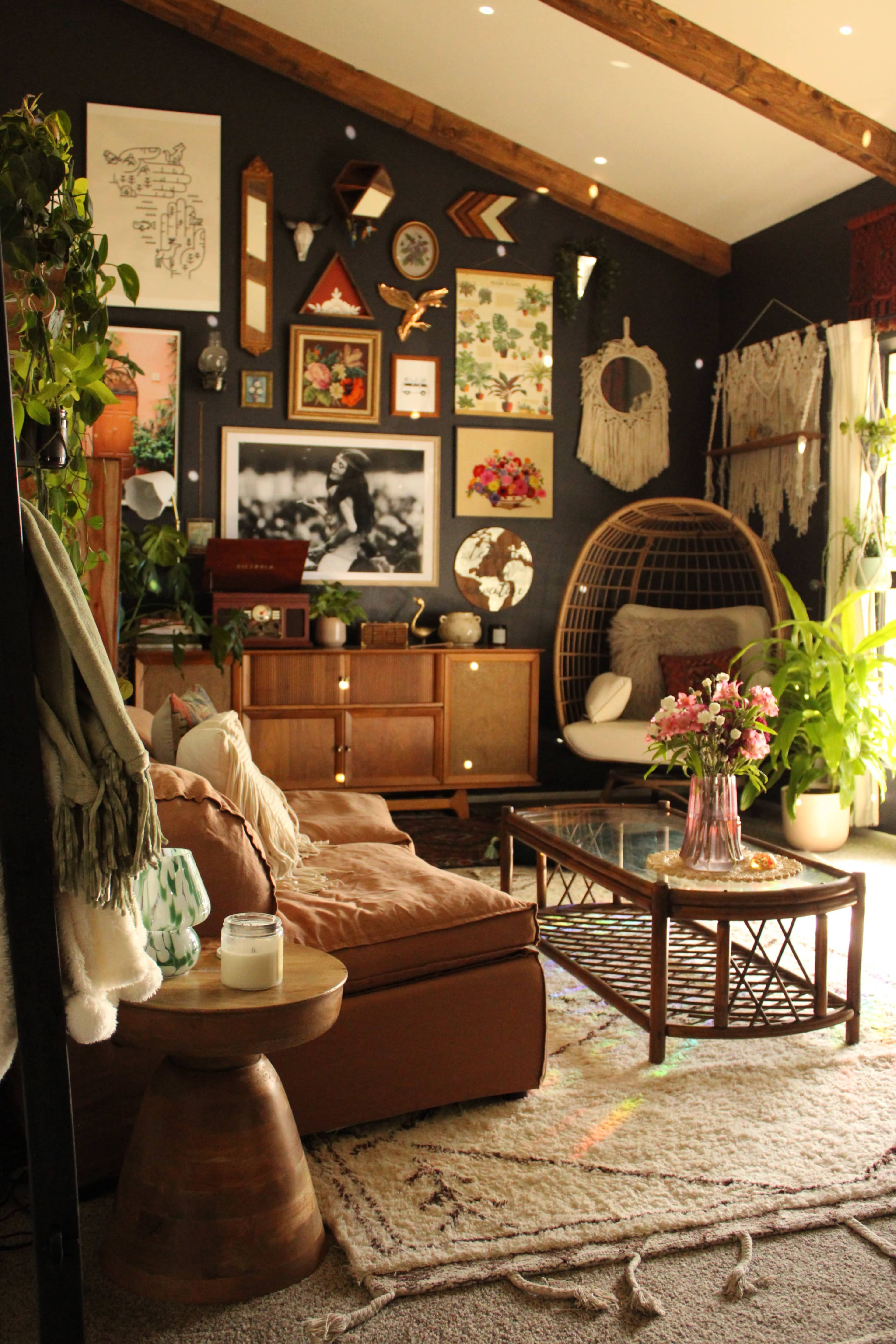 A cozy living room with a dark wall, a mix of decorative plants, art pieces, and comfortable furniture, including a rattan chair and a wooden coffee table.