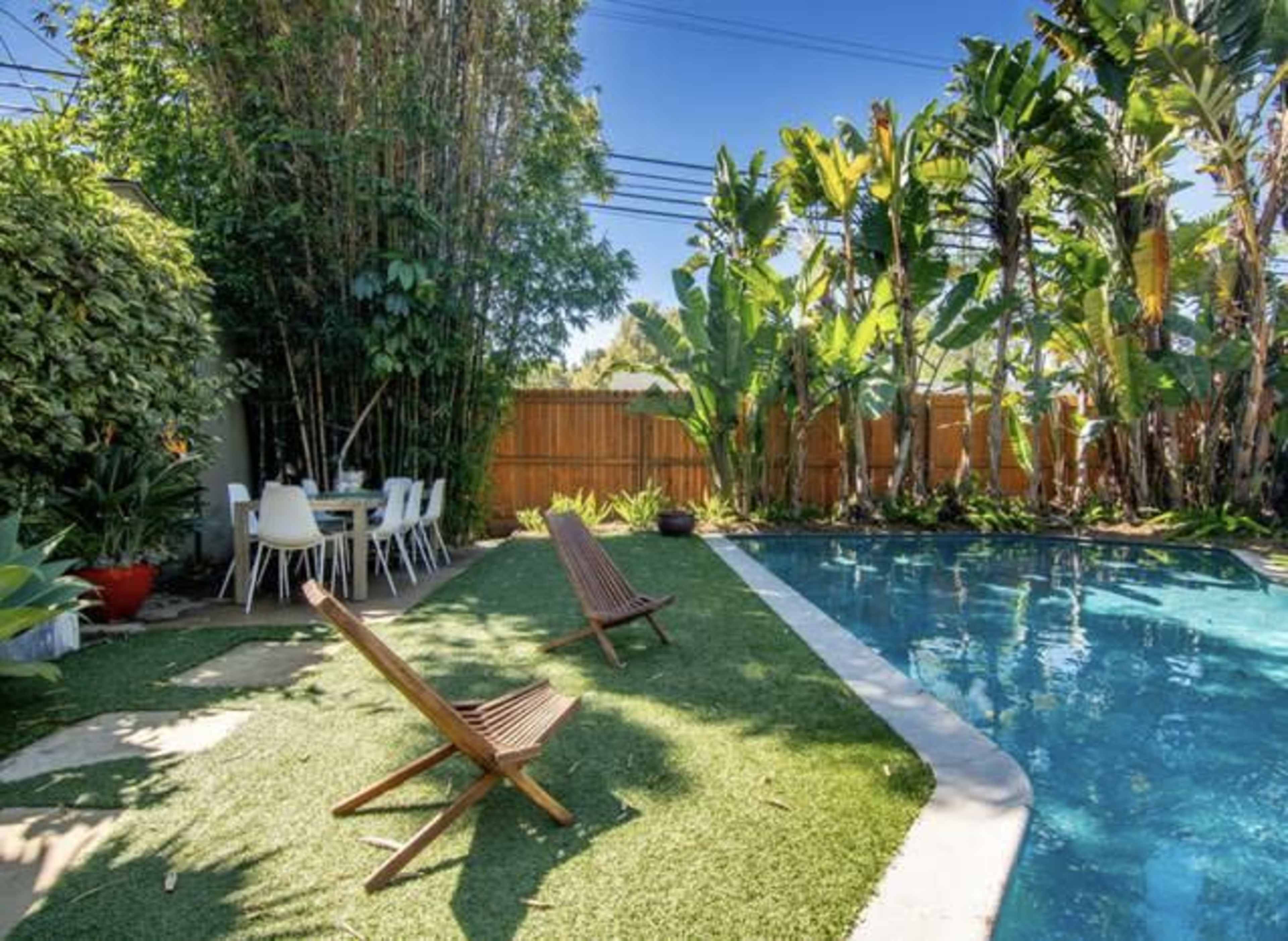 A swimming pool surrounded by tropical plants and a seating area with a table and chairs in a backyard.