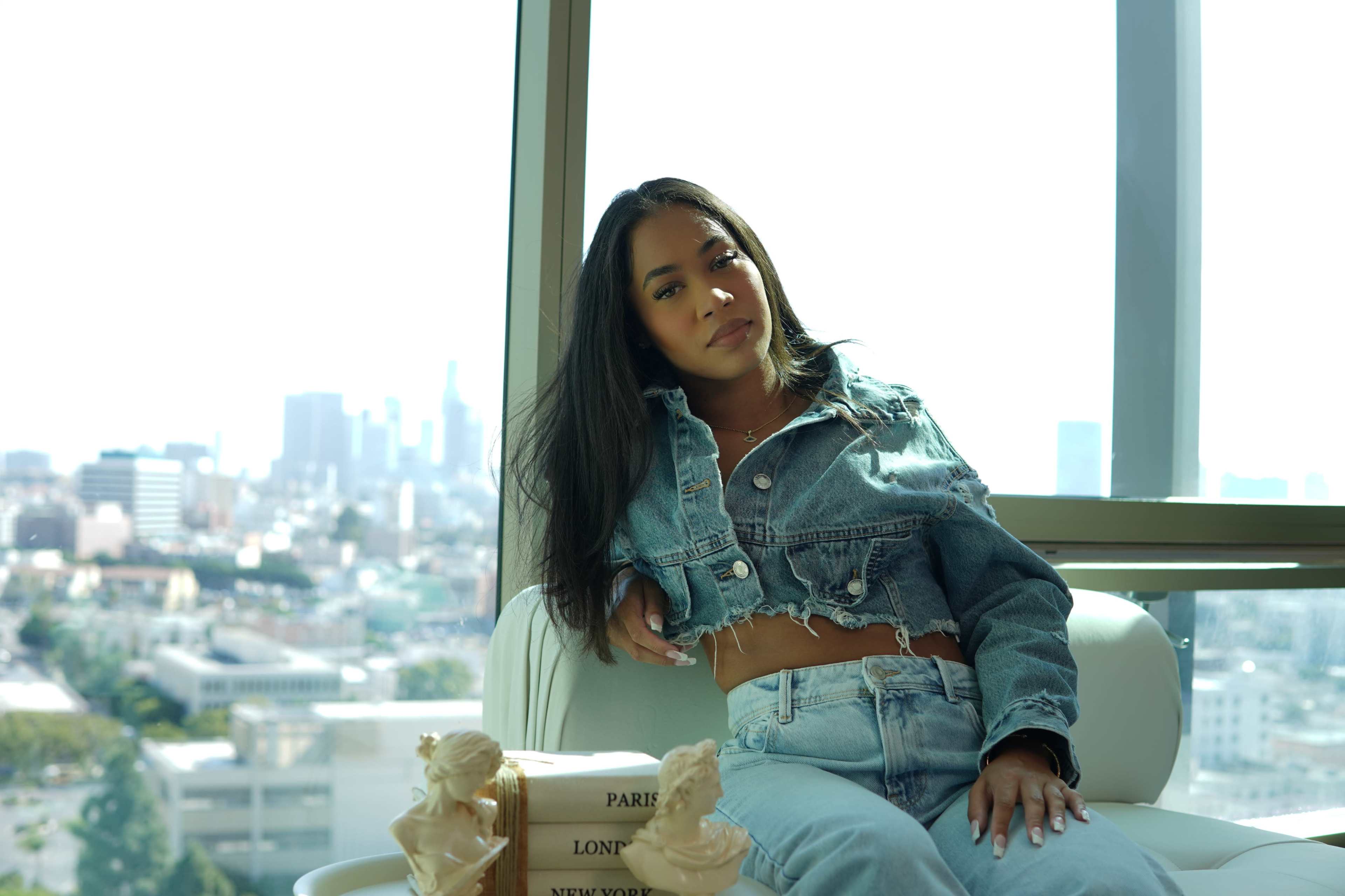 A woman in a denim jacket and high-waisted jeans sits in a chair with a city skyline view behind her.