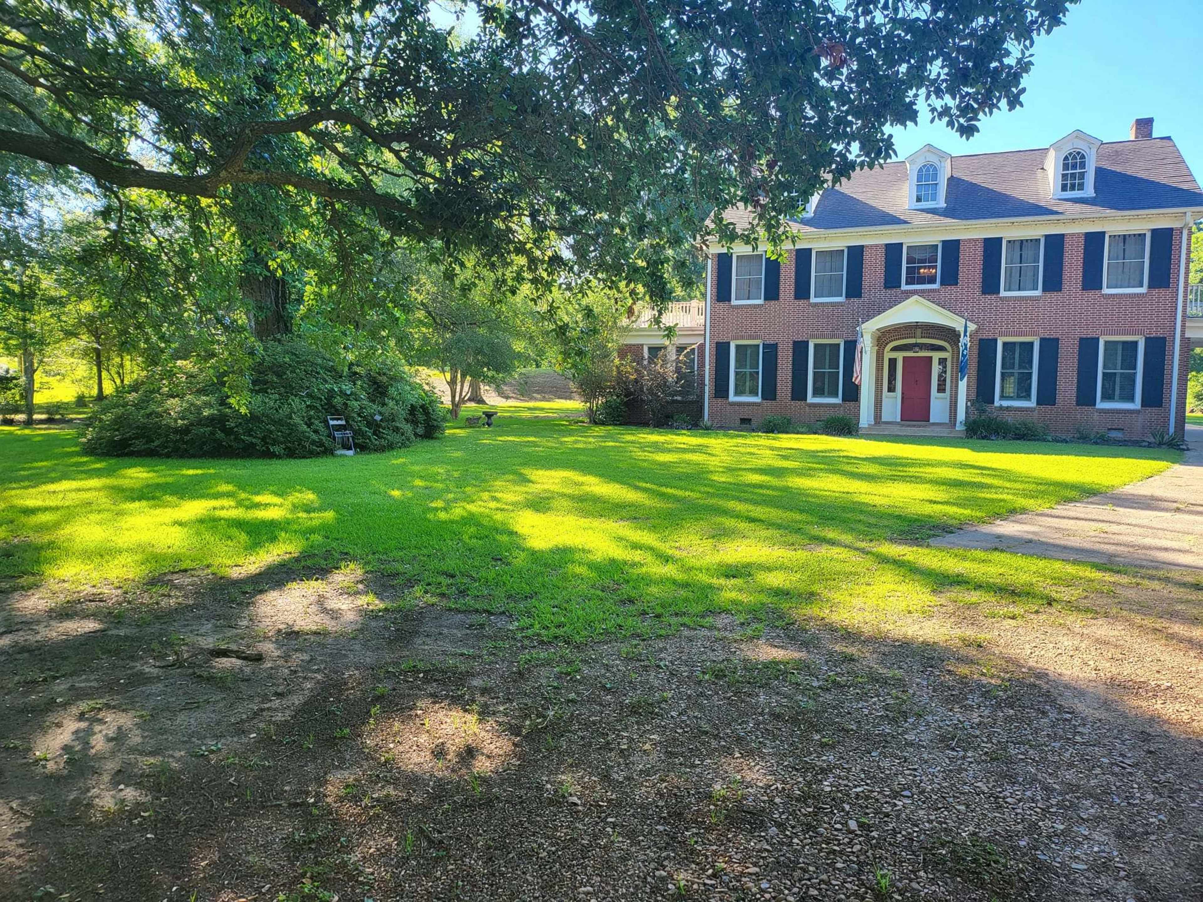 A large brick house with white trim and a red door sits in a grassy yard surrounded by trees.