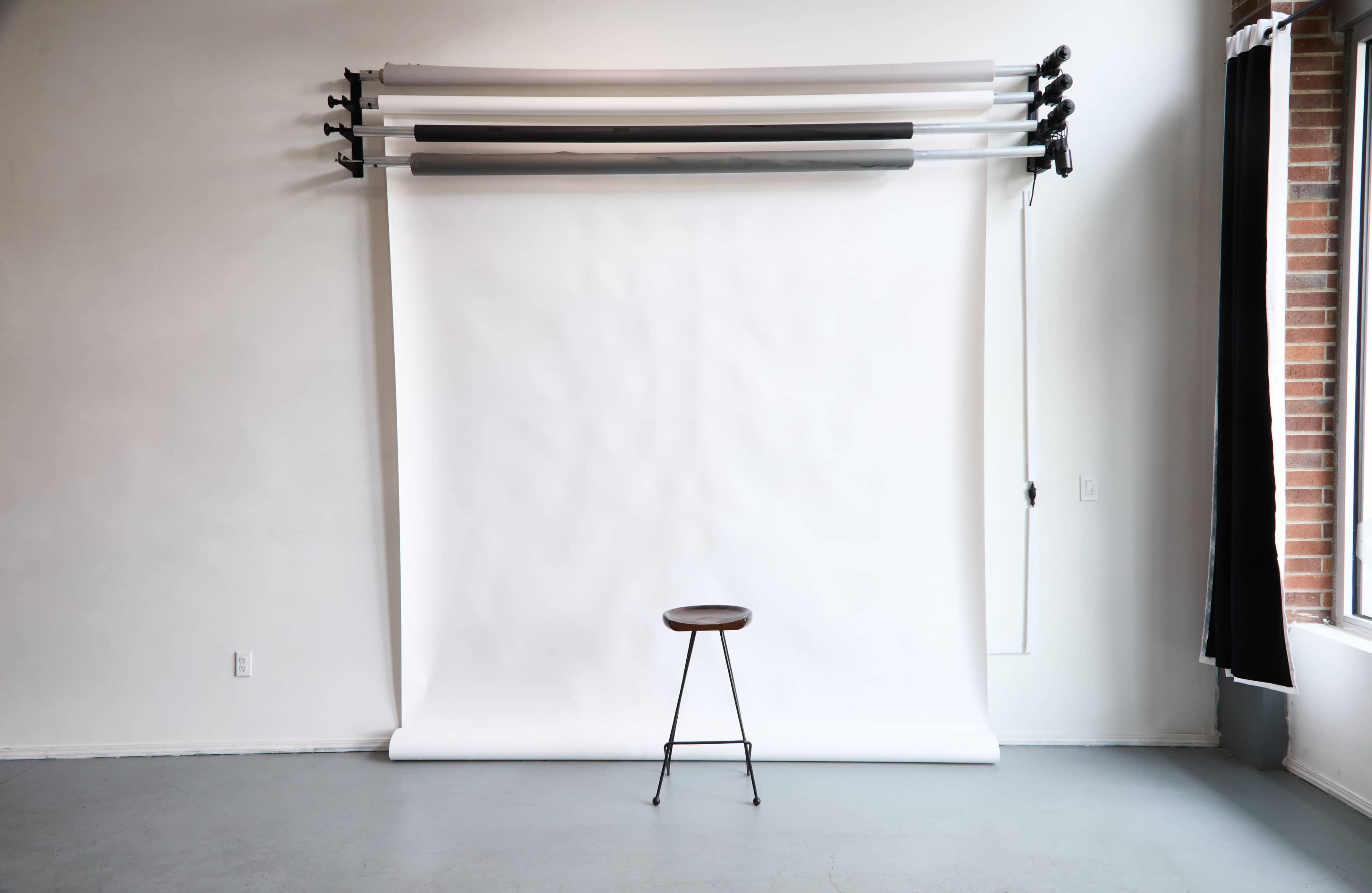 The image shows a minimalist studio setup with a white backdrop and a single wooden stool positioned in front of it.