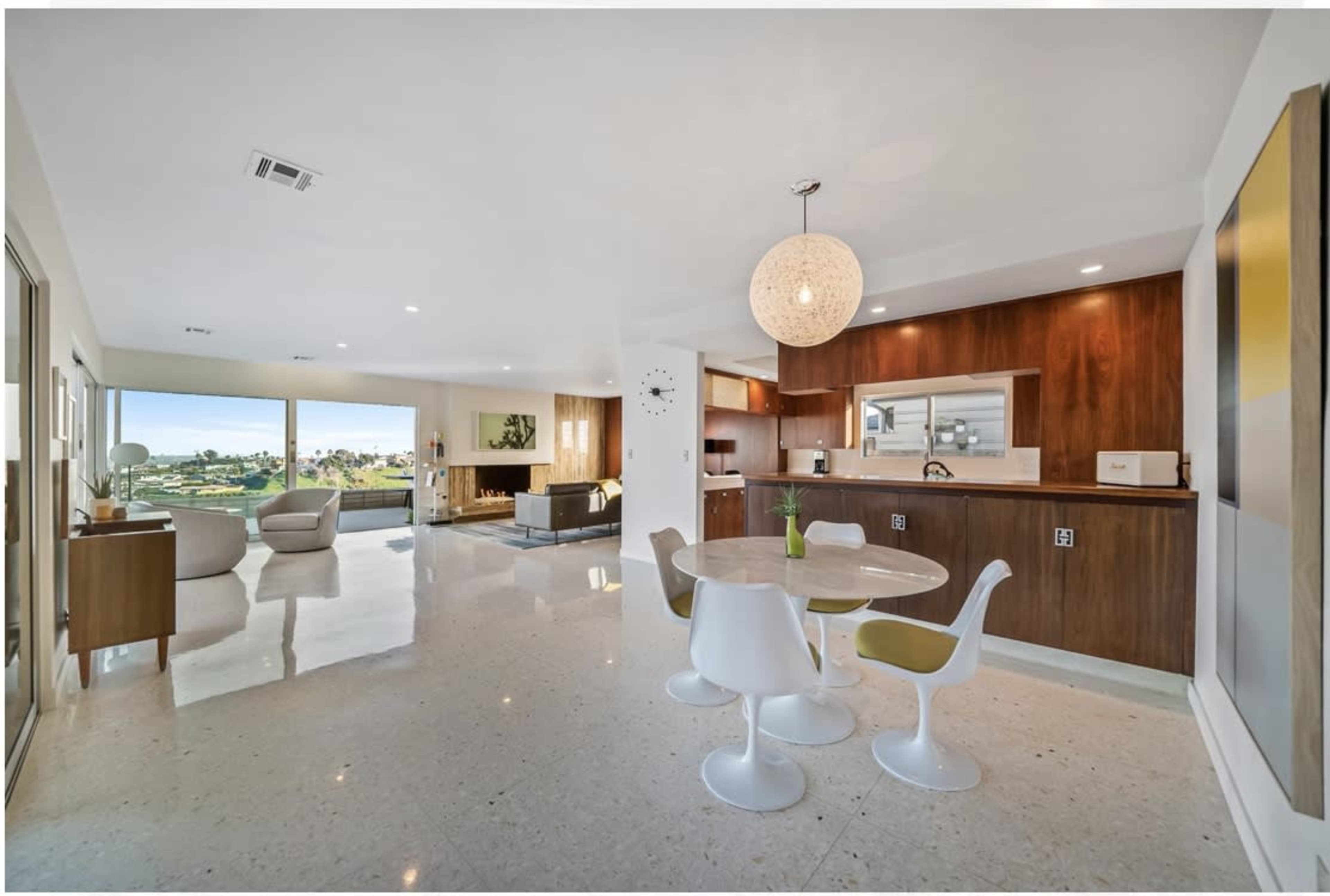 A modern living space with a dining area featuring a round table and chairs, a kitchenette, and large windows overlooking a landscape.