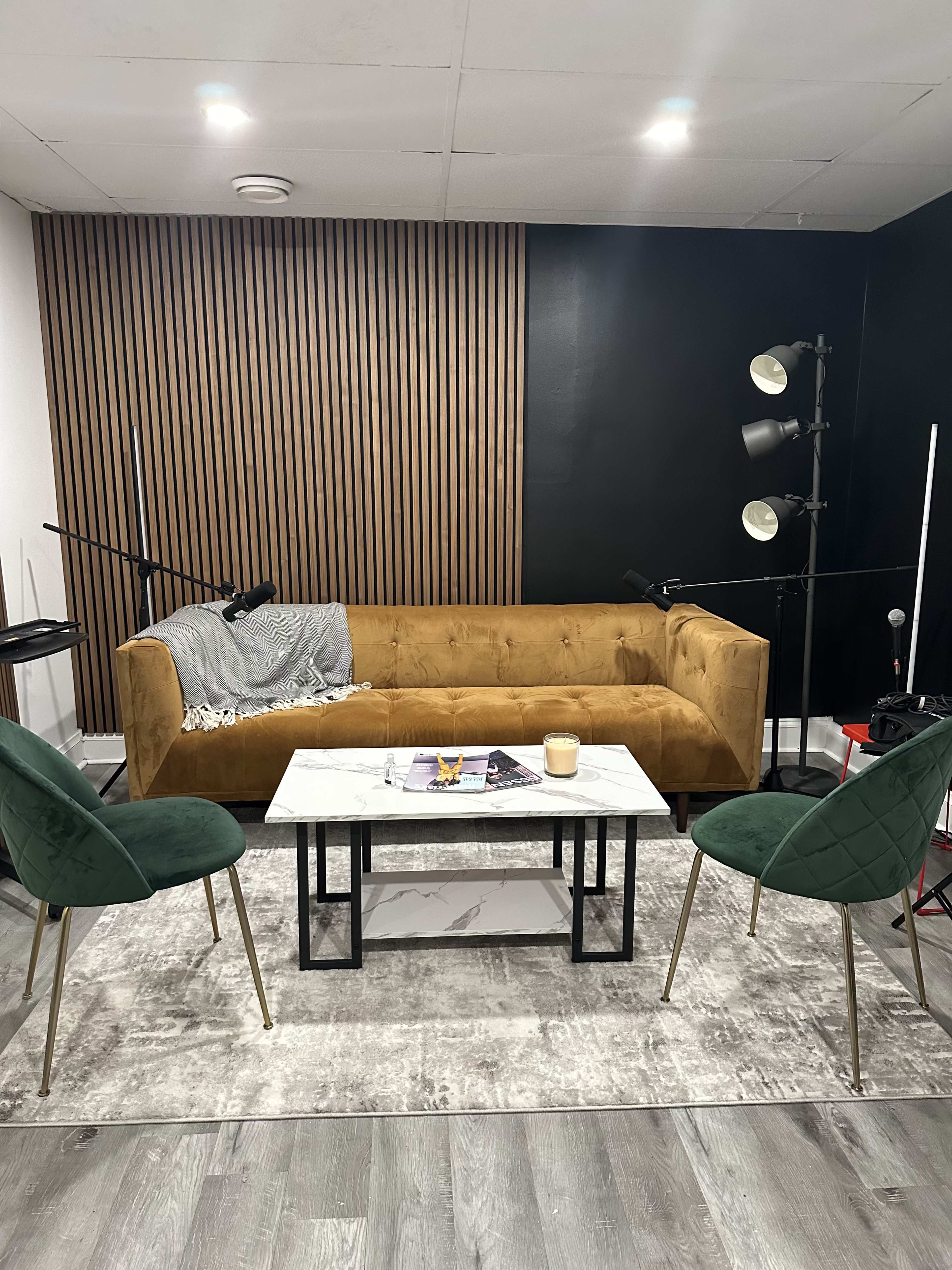 A living room setup with a brown velvet sofa, a marble coffee table, and two green chairs, all set against a backdrop of black and wooden paneling.