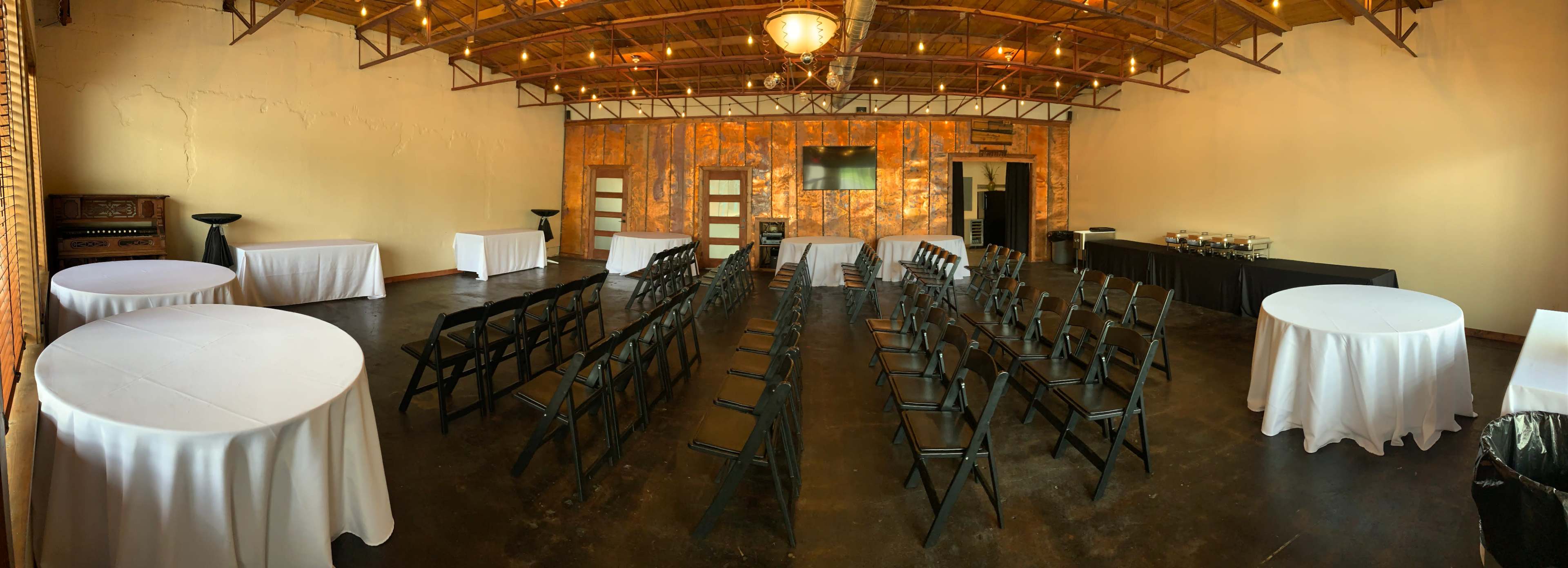 A spacious, well-lit event room set up with black chairs arranged in rows, round tables covered in white linens, and a buffet table along one wall.